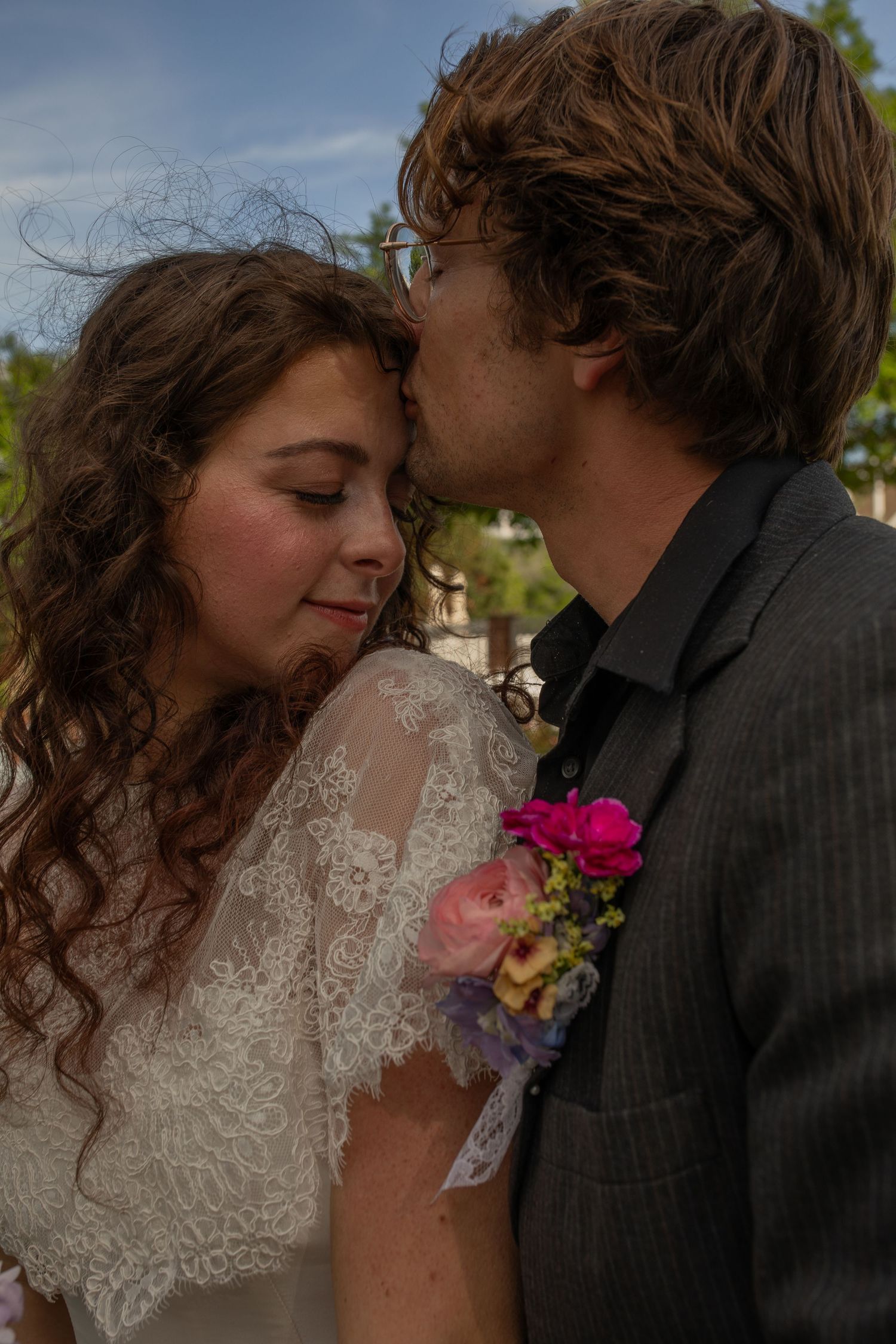 Groom in gray suit with floral boutonniere kissing bride in white lace dress on their wedding day outdoors.