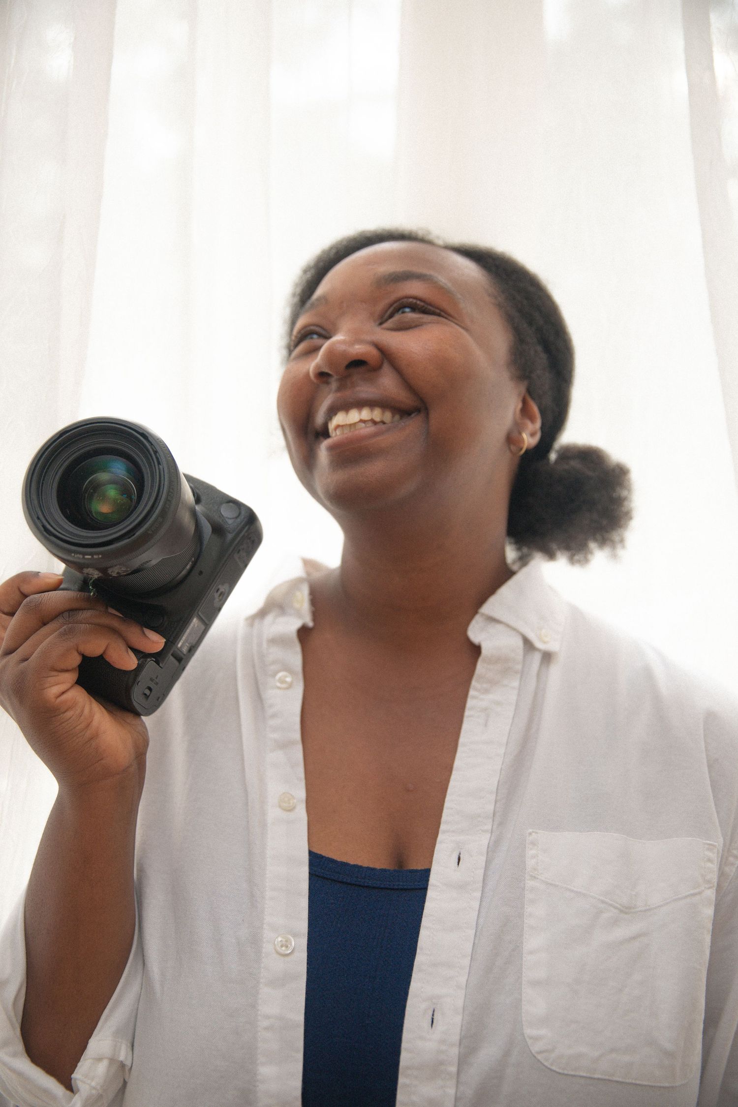 Smiling woman holding a DSLR camera, wearing a white shirt over a blue top, standing near a bright window.