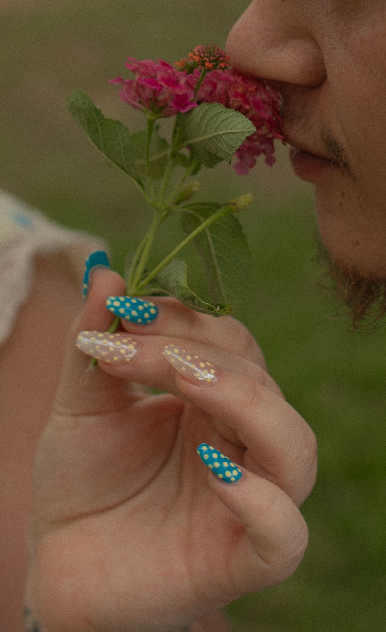 Close-up of a person smelling pink flowers, showcasing decorative teal and white polka dot nail art on long nails.