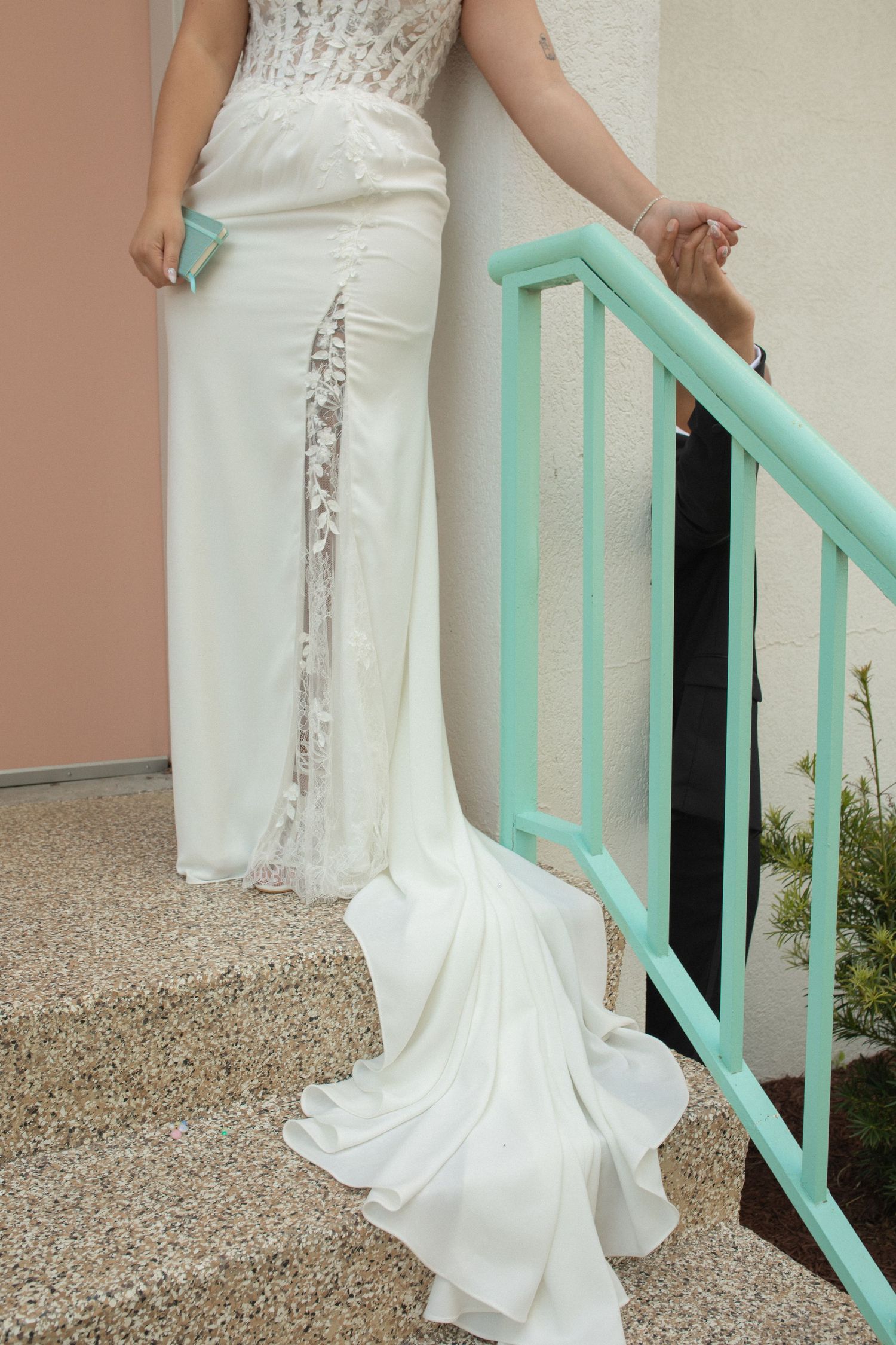 Bride in elegant white wedding gown with lace detail and train standing on steps with mint green railing.