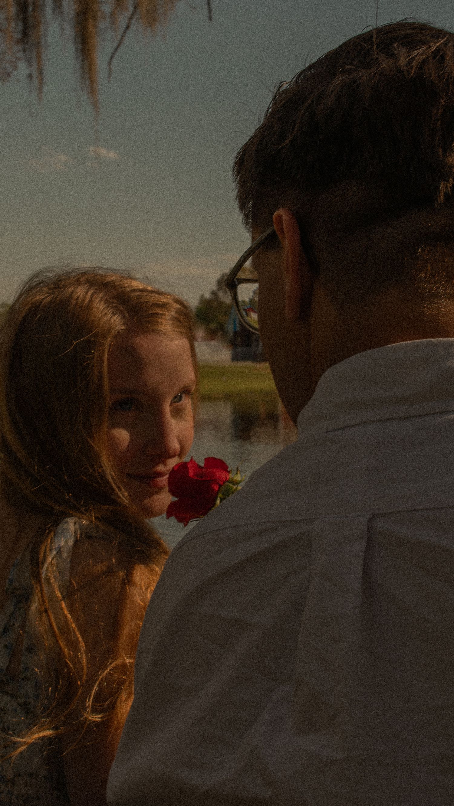 A romantic moment captured at sunset by a lake with a red rose between two people.