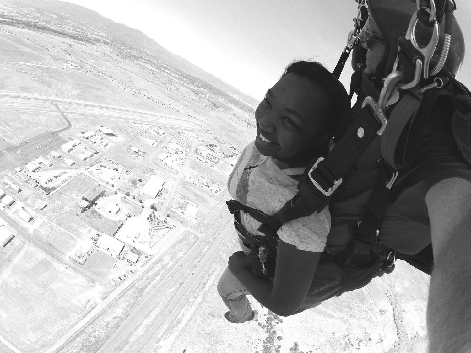Black and white photo of a skydiver in tandem harness smiling during freefall over desert landscape.