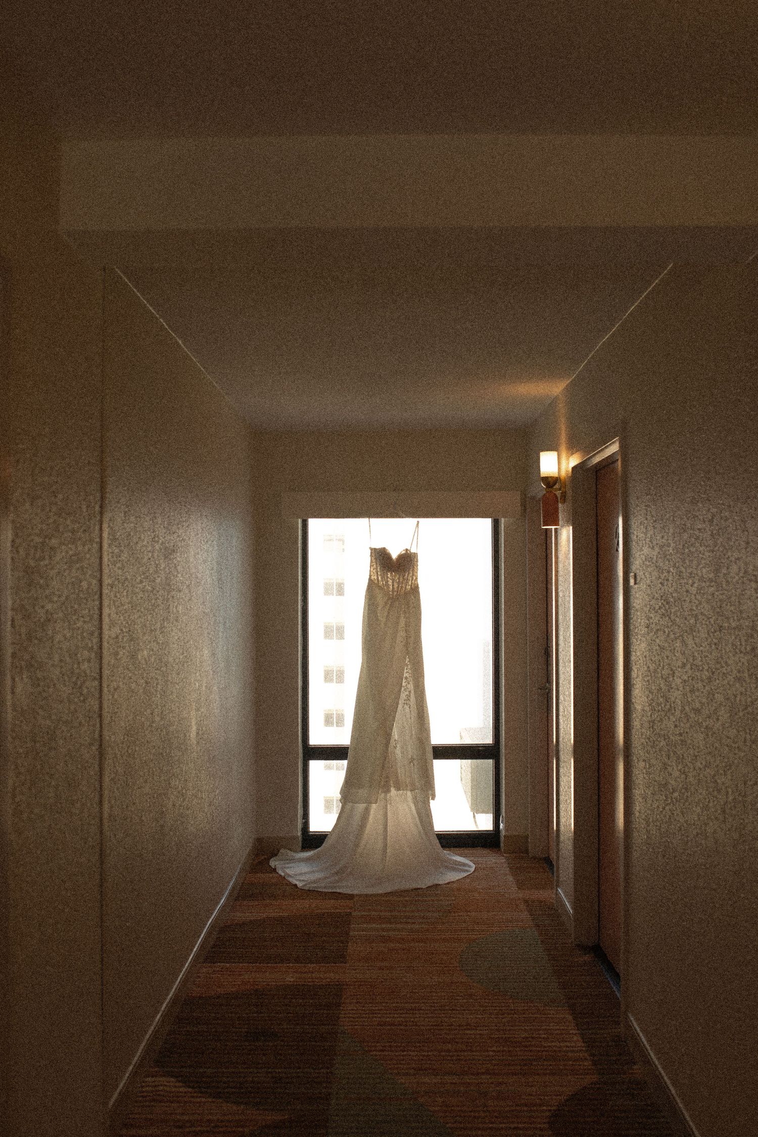 A white wedding dress hanging in a dimly lit hotel hallway corridor with a bright window in the background.