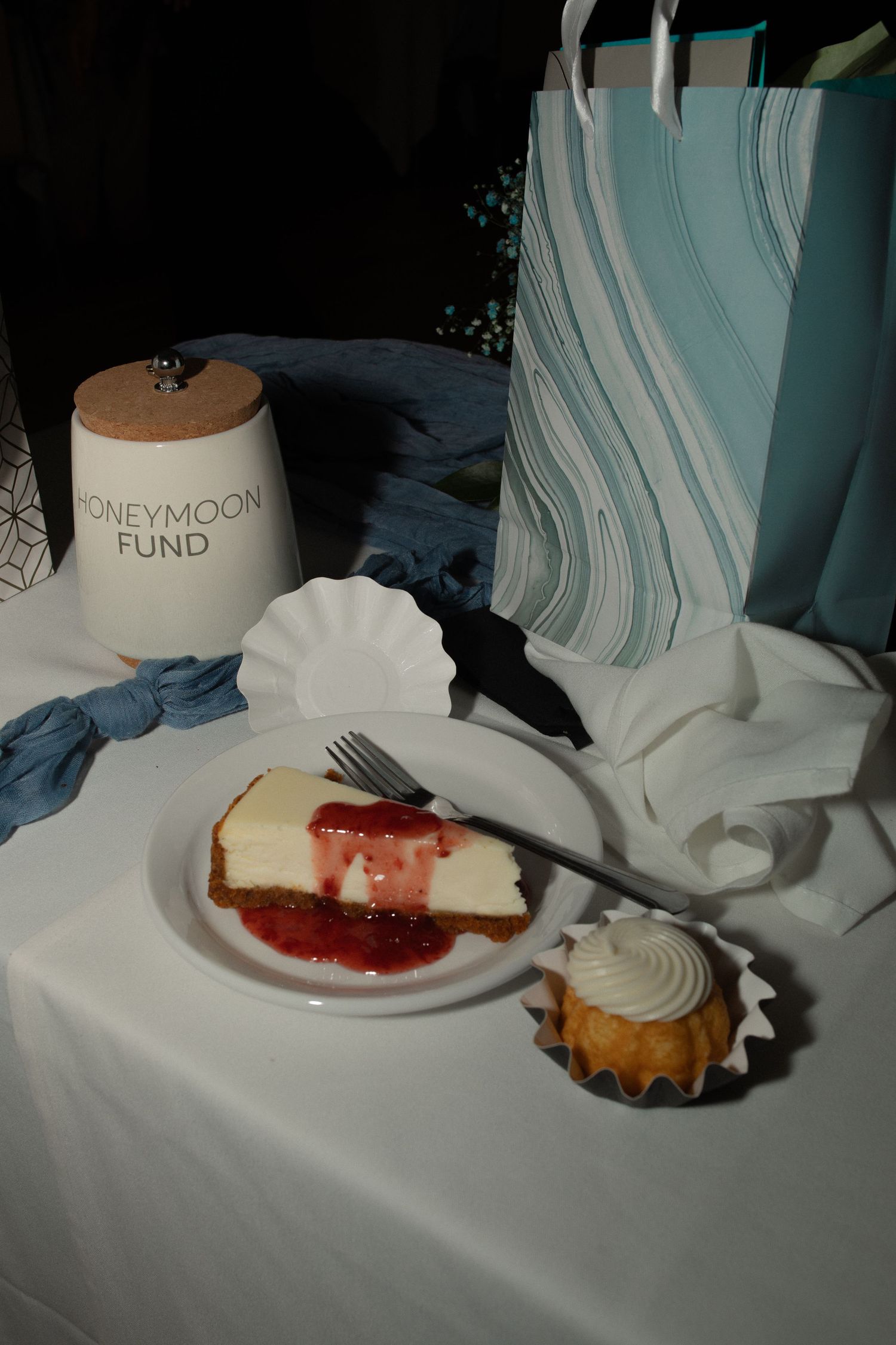 Wedding dessert table with cheesecake, cupcake, honeymoon fund jar, and teal gift bag on white tablecloth.
