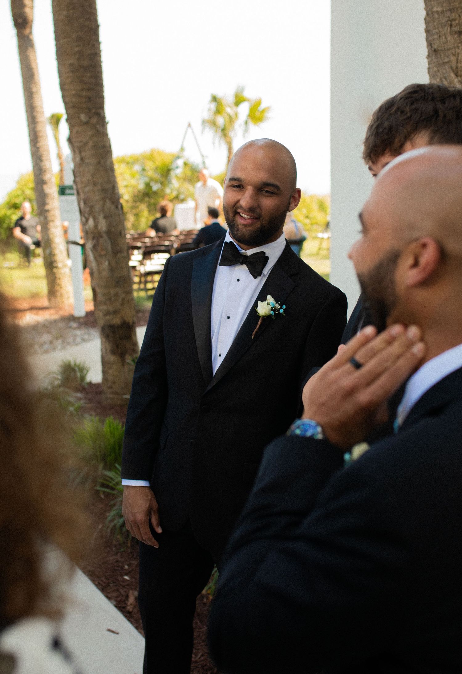 Groom in black tuxedo with boutonniere smiling at wedding ceremony outdoors near palm trees.