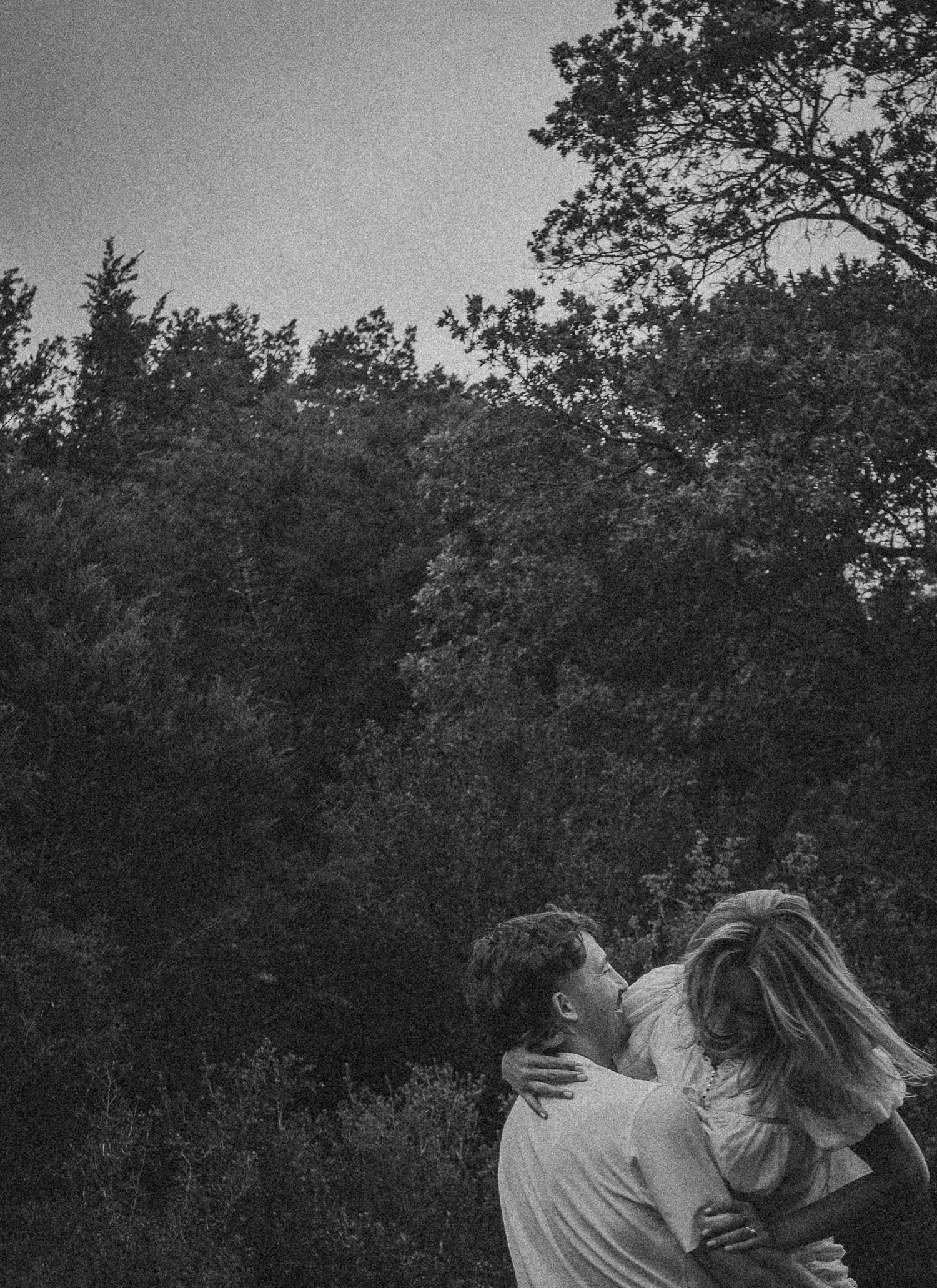 Black and white photo of a couple embracing and kissing outdoors surrounded by lush trees in a romantic forest setting.
