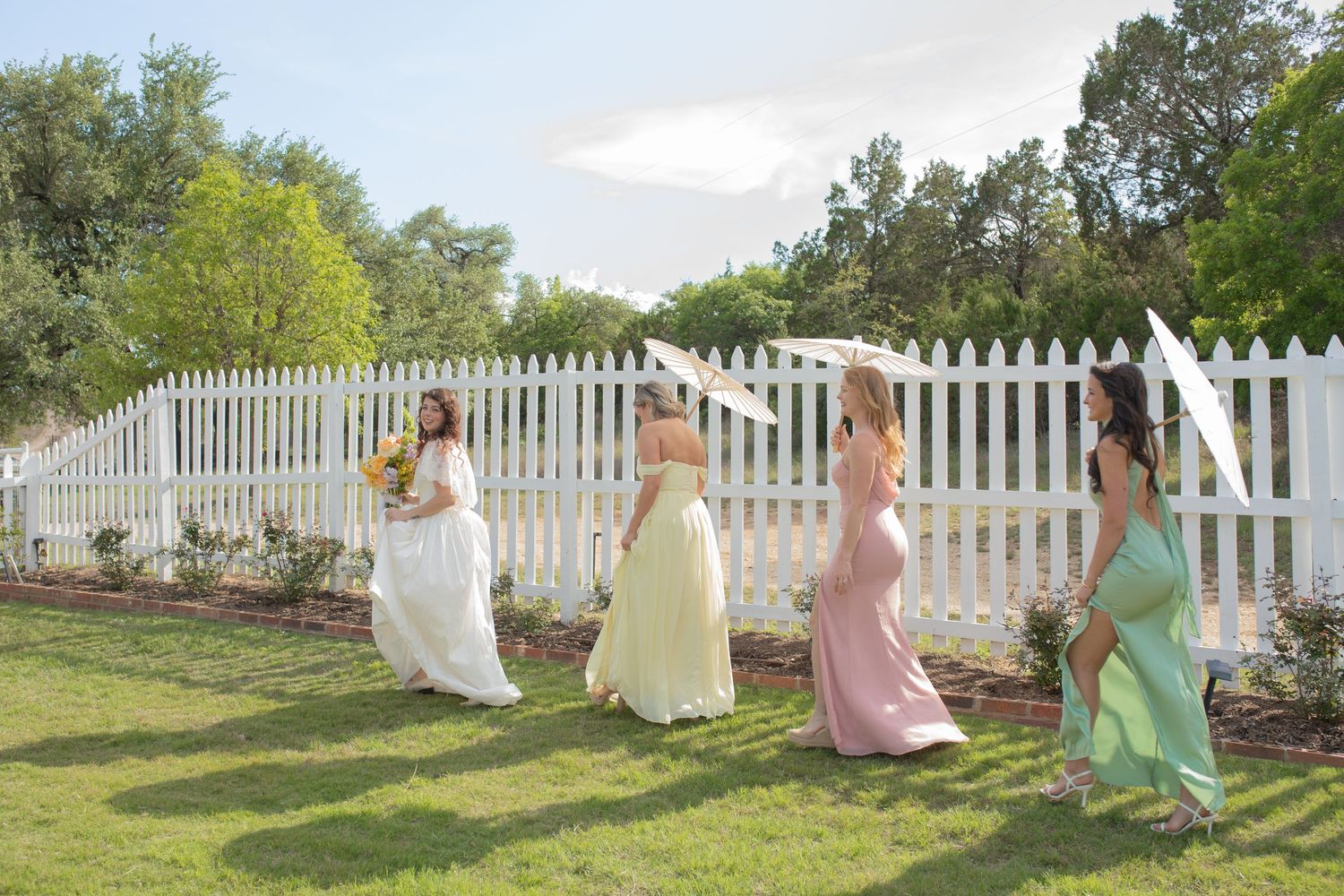 Bride and bridesmaids in colorful gowns walking on green lawn beside white picket fence outdoors.