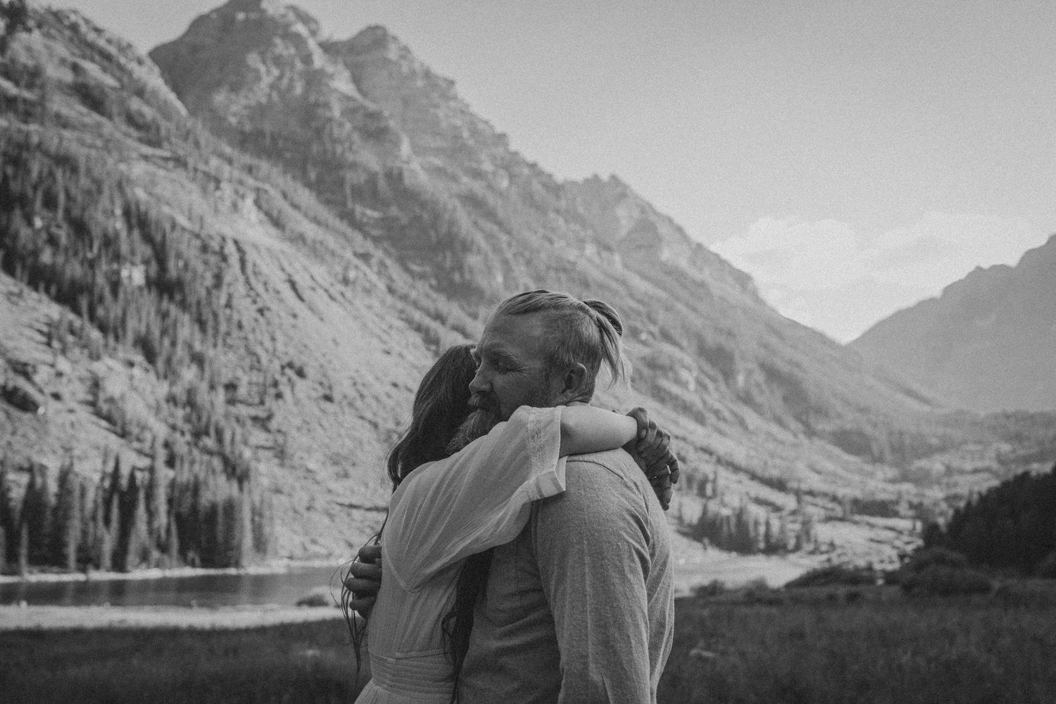 Black and white photo of a couple embracing tenderly with mountains and a lake in the background.