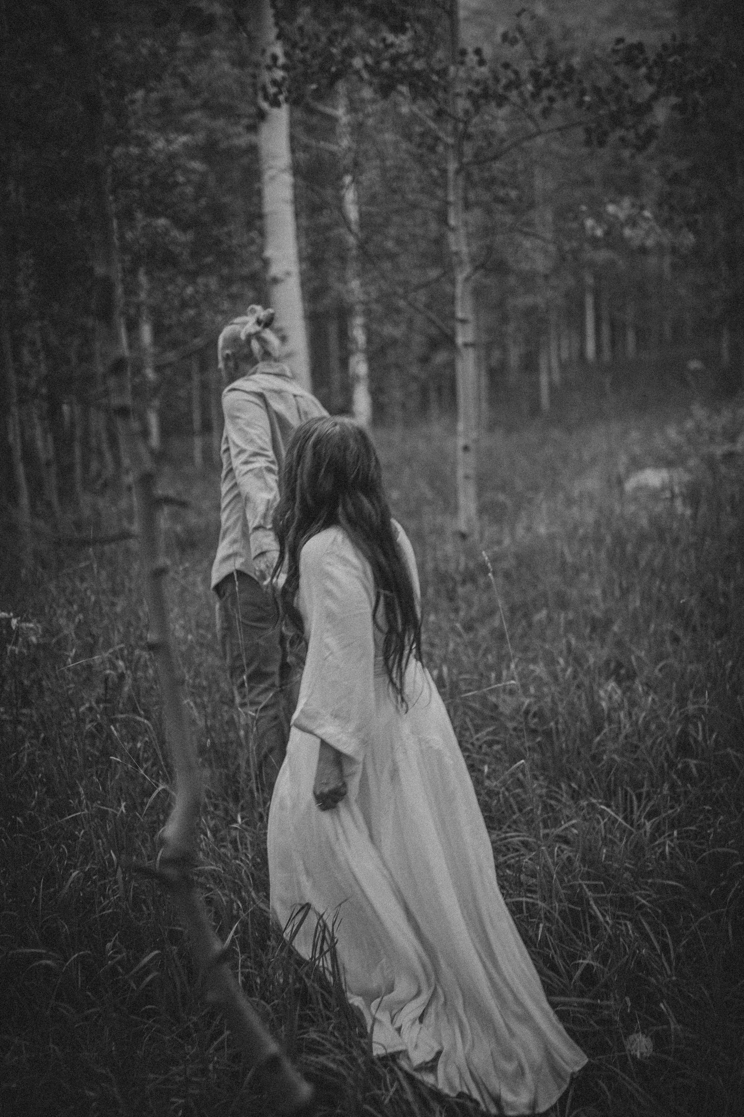 Black and white photo of a couple walking hand in hand through a misty forest, woman in flowing white dress.