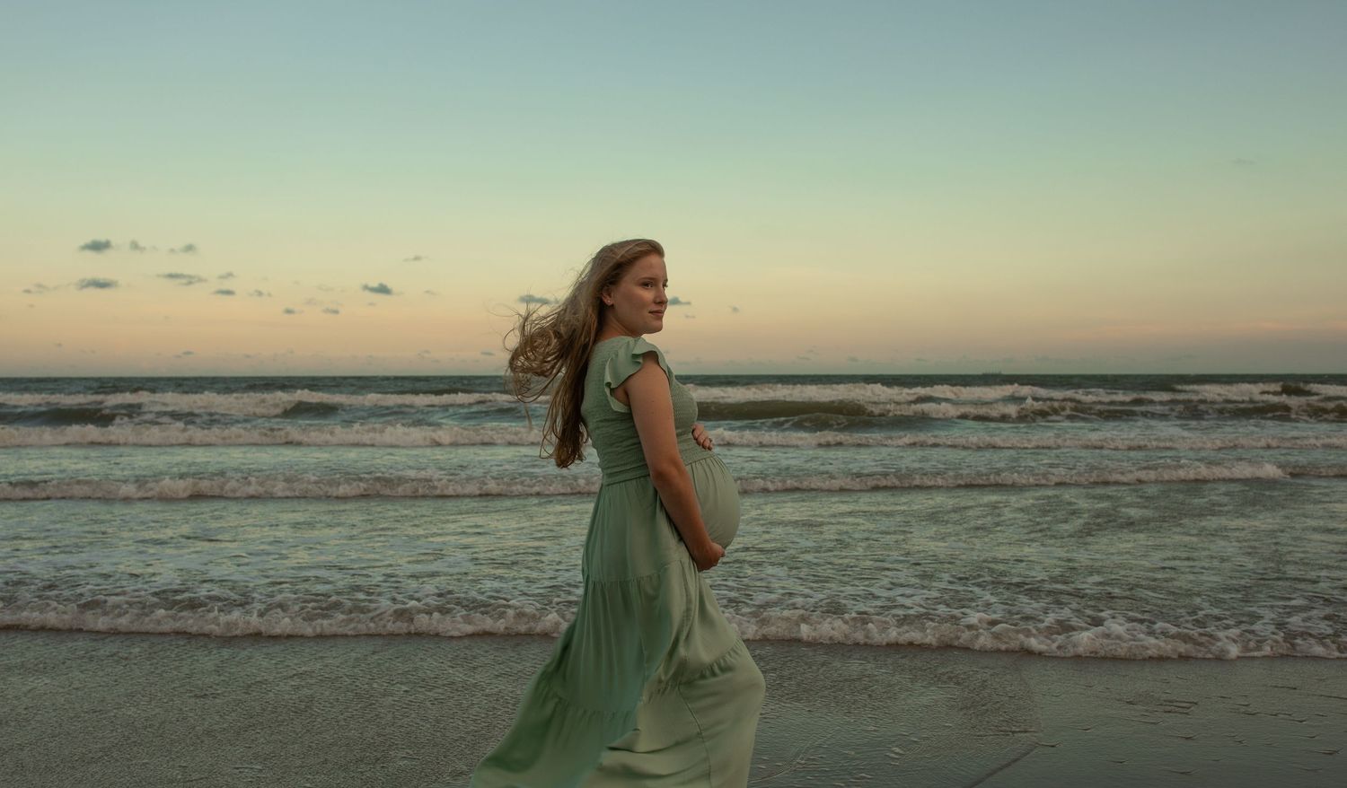 A figure in a flowing mint green dress walks along a beach at sunset as waves roll in from the ocean.