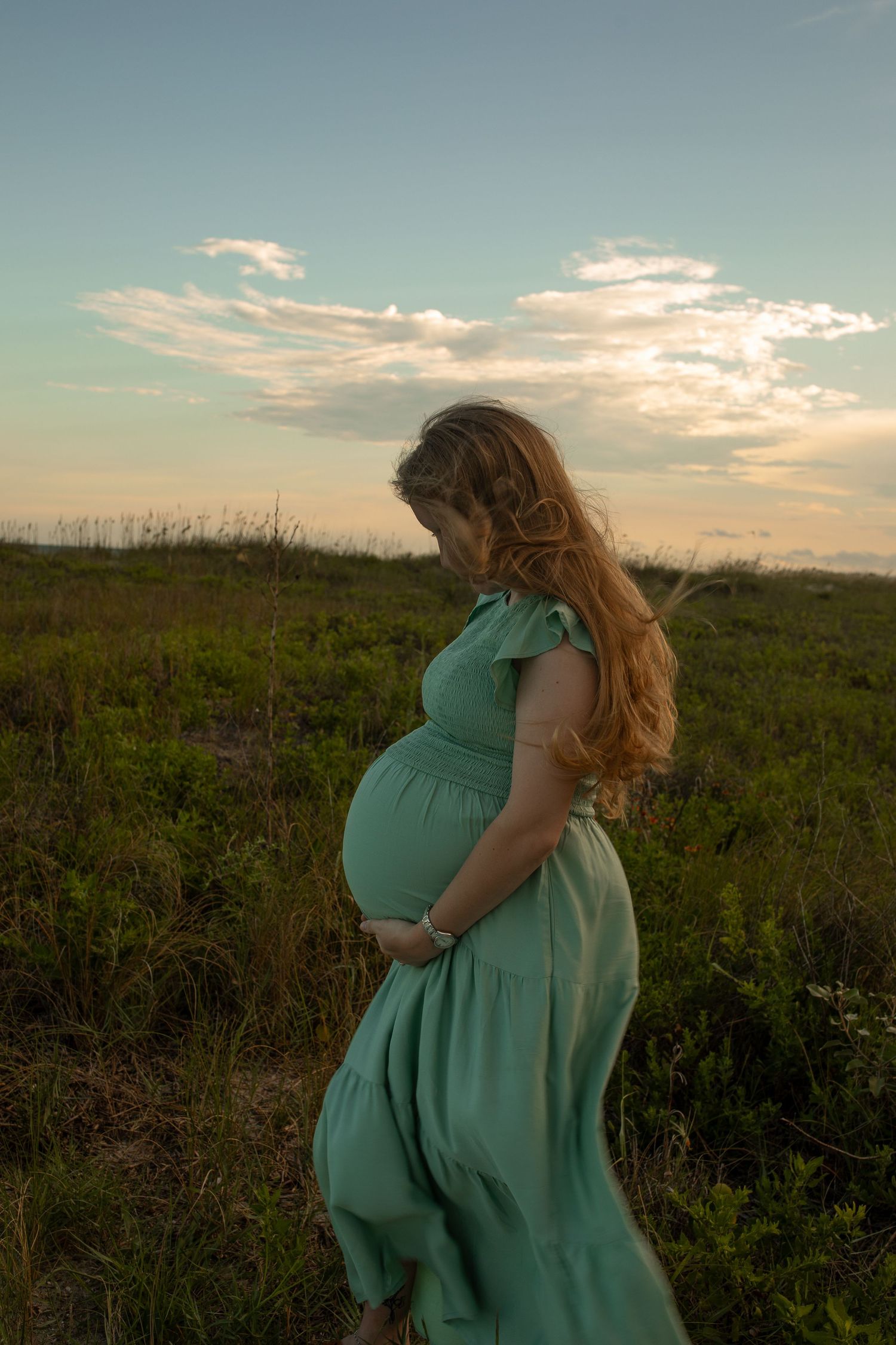 A flowing mint green dress catches the wind in a coastal field at sunset.