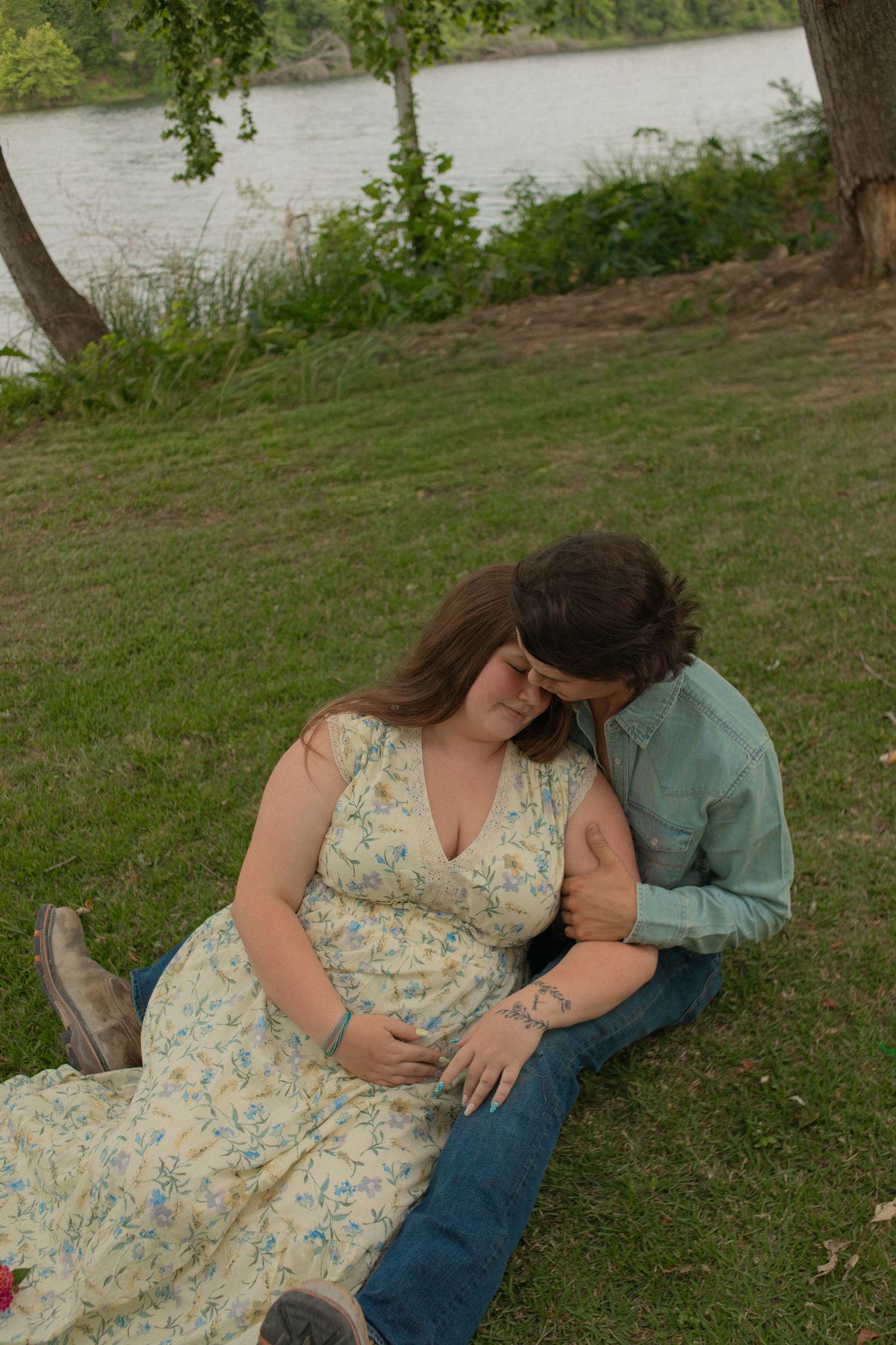 Pregnant couple sitting on grass by a river, woman in floral dress with partner embracing her baby bump outdoors.
