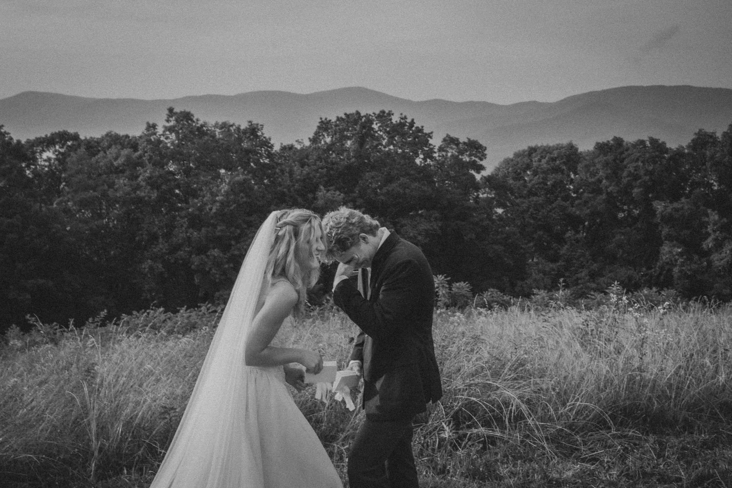 Black and white photo of a bride and groom sharing an intimate moment in a field with misty mountains in the background.