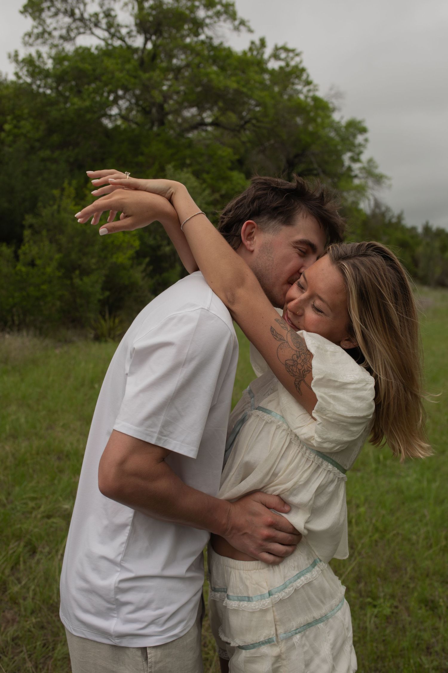 A couple embraces romantically in a lush green field, the man nuzzling the smiling woman in a white dress.