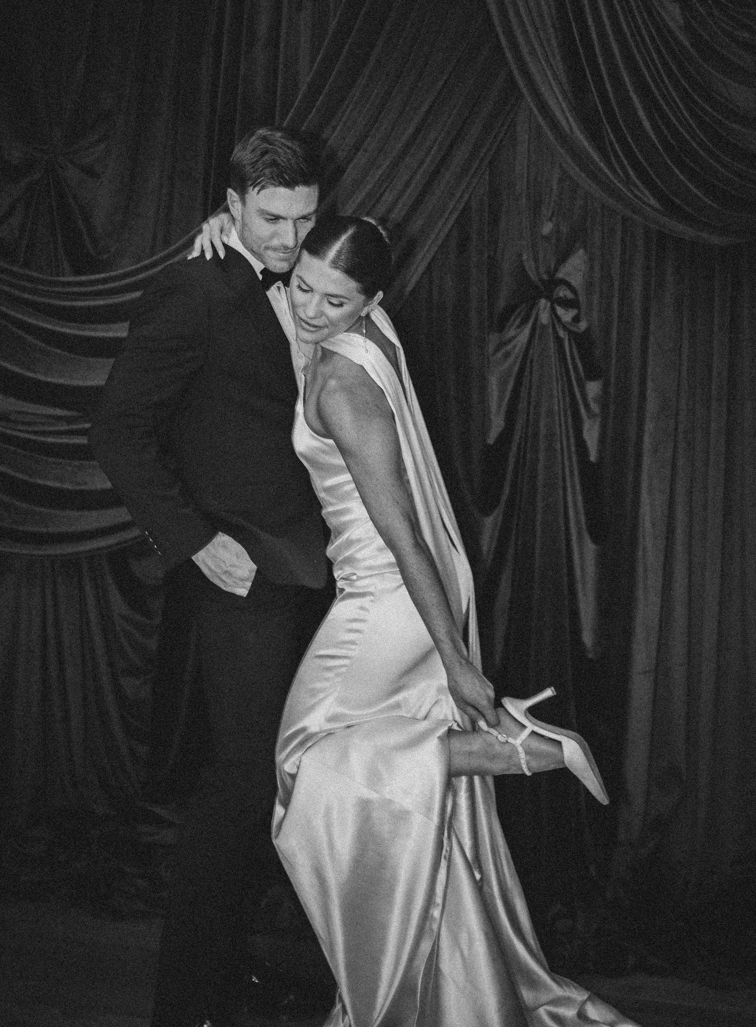 Elegant couple in black and white photo, woman in satin gown adjusting heel, man in tuxedo, dramatic curtain backdrop.