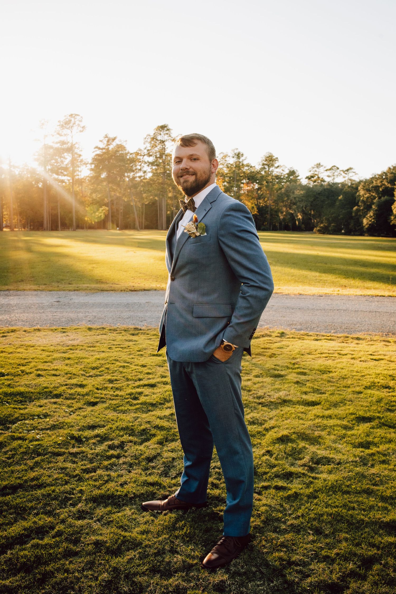 A person in a blue suit stands on grass at sunset with golden sunlight filtering through trees in the background.