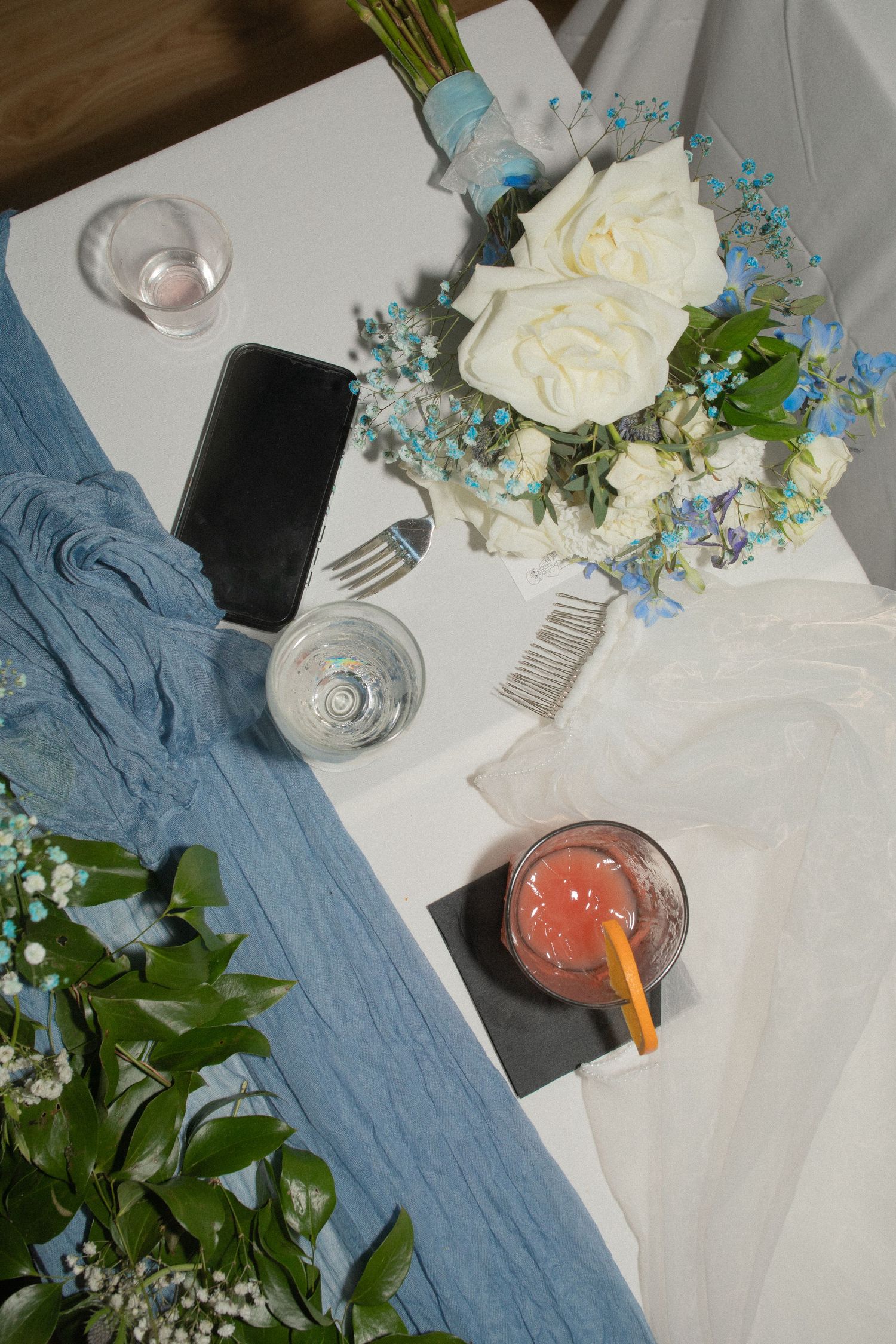 White roses and blue flowers bouquet on table with phone, glasses, and pink drink at a wedding reception.