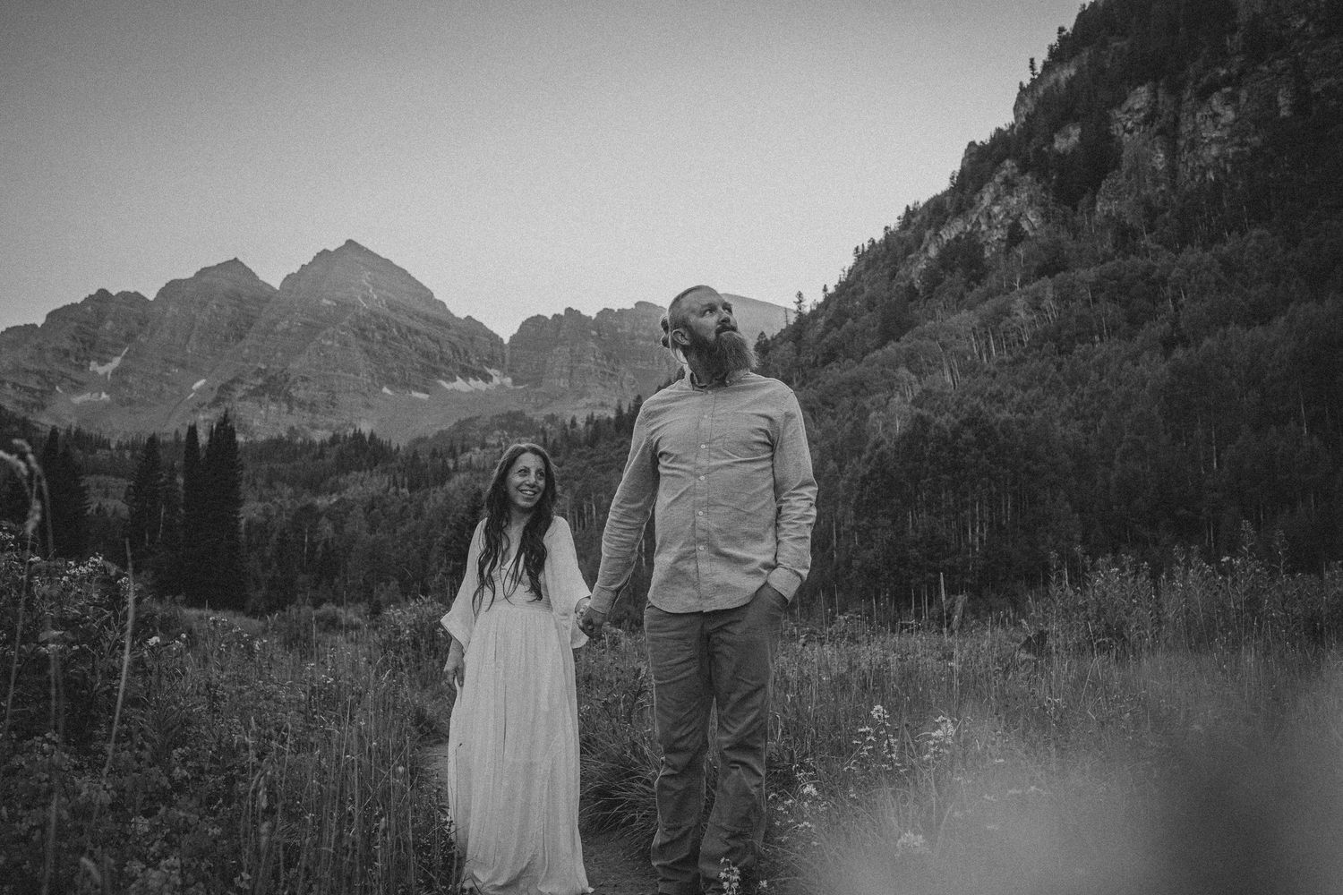 Black and white photo of a couple holding hands in a mountain meadow, woman in white dress, dramatic peaks behind them.