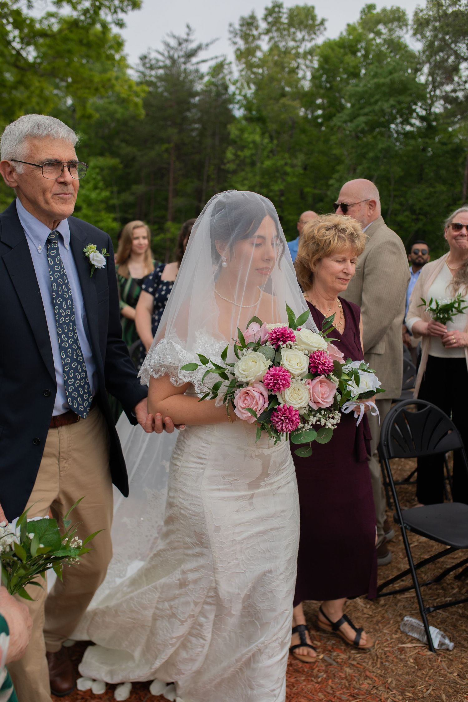 Bride in white lace gown and veil walks down aisle with parents, holding pink and white bouquet at outdoor wedding.