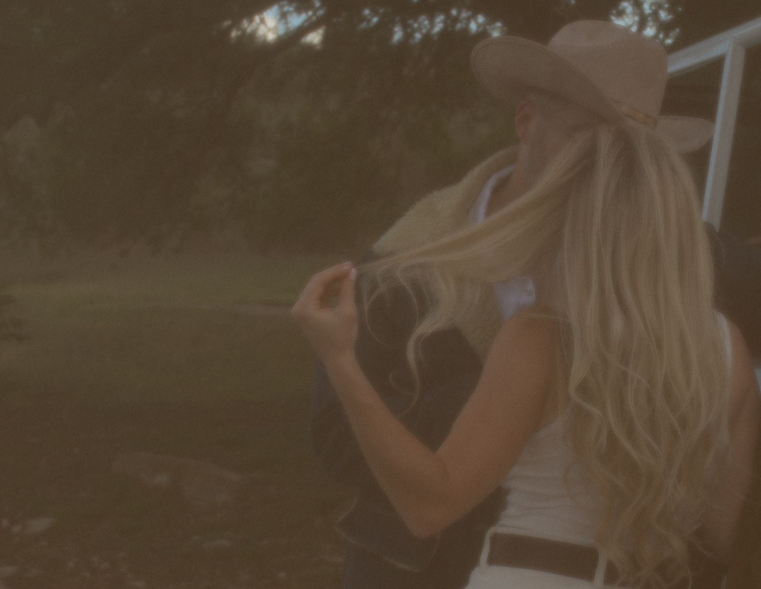 Blonde woman in cowboy hat with long hair standing outdoors near a vehicle at golden hour.