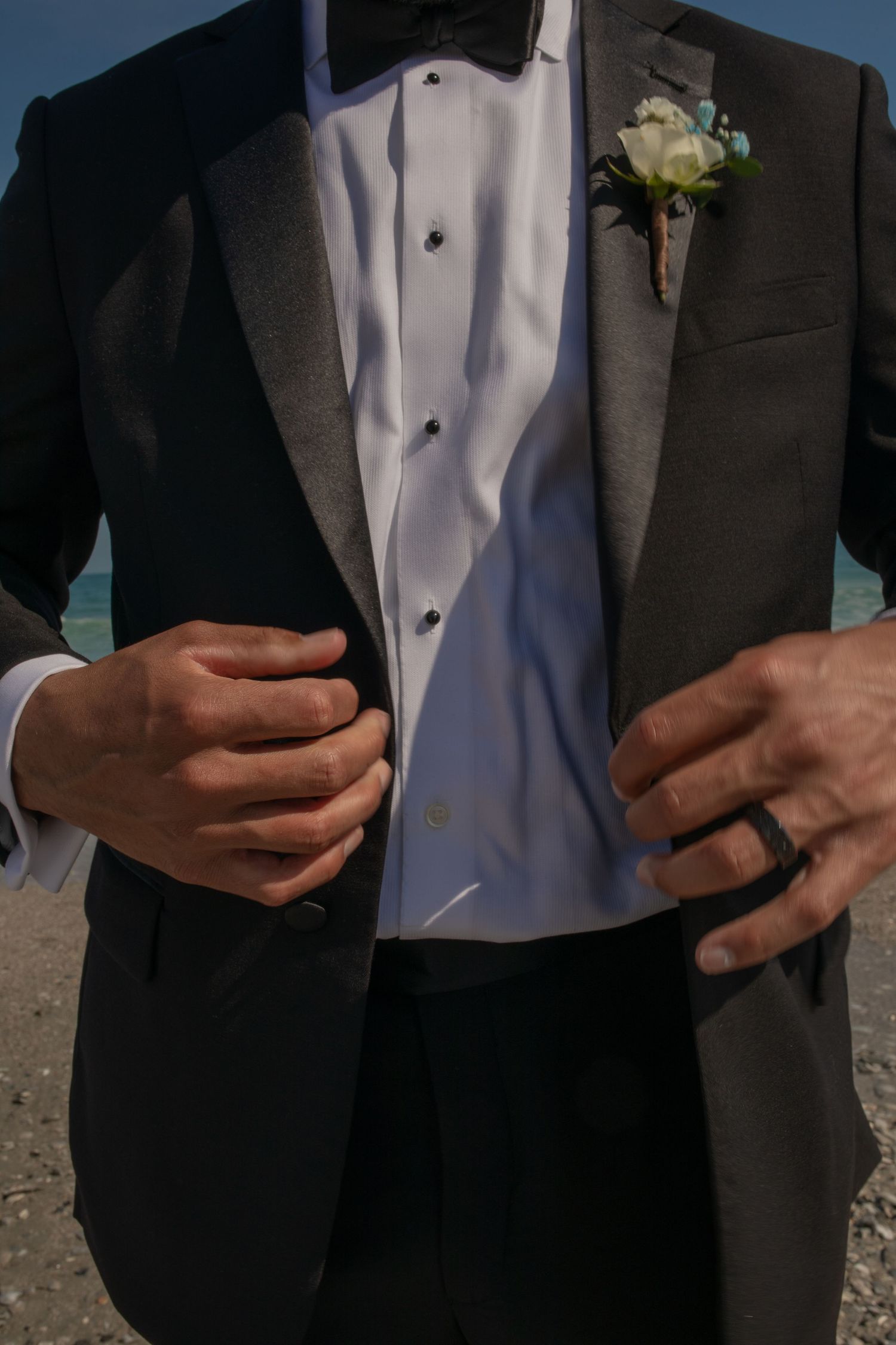 Groom in black tuxedo with white boutonniere adjusting jacket lapels on beach wedding day.