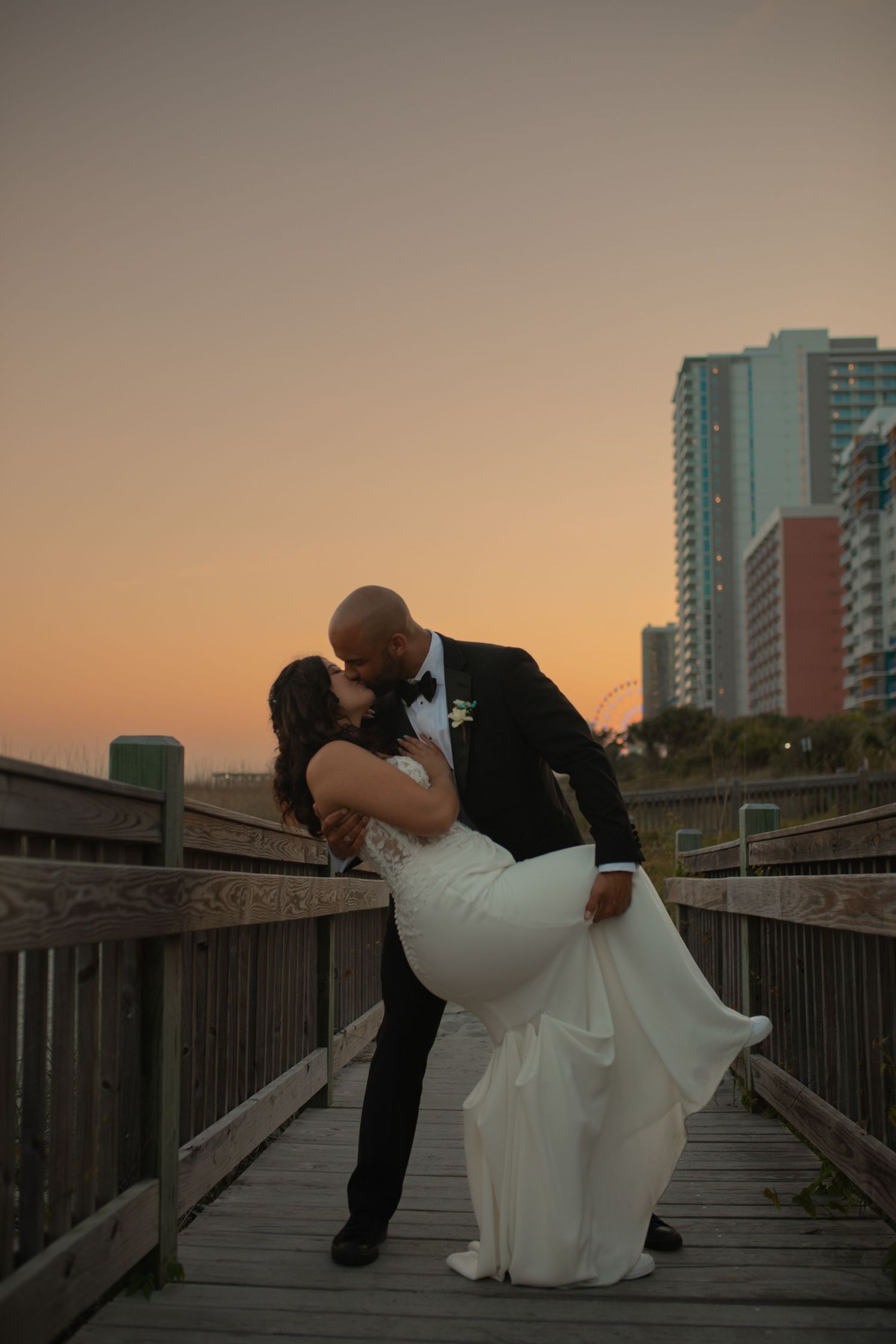 Bride and groom share a romantic dip kiss on a boardwalk at sunset with city skyline in background.