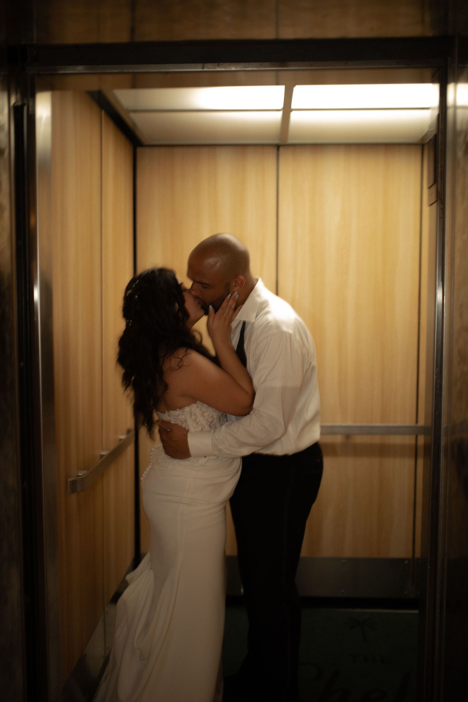 Newlywed couple sharing a romantic kiss in an elevator, bride in white gown and groom in white shirt.