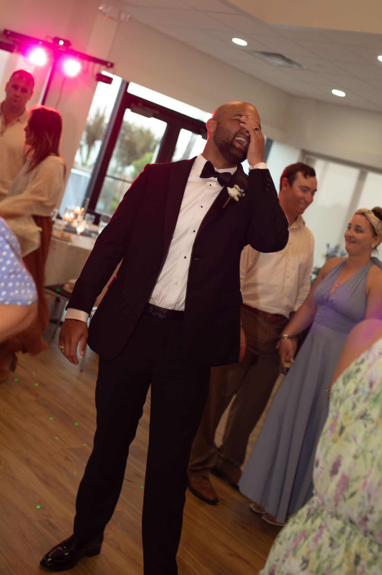 A groom in a dark tuxedo with bow tie dances at his wedding reception while guests watch and smile behind him.