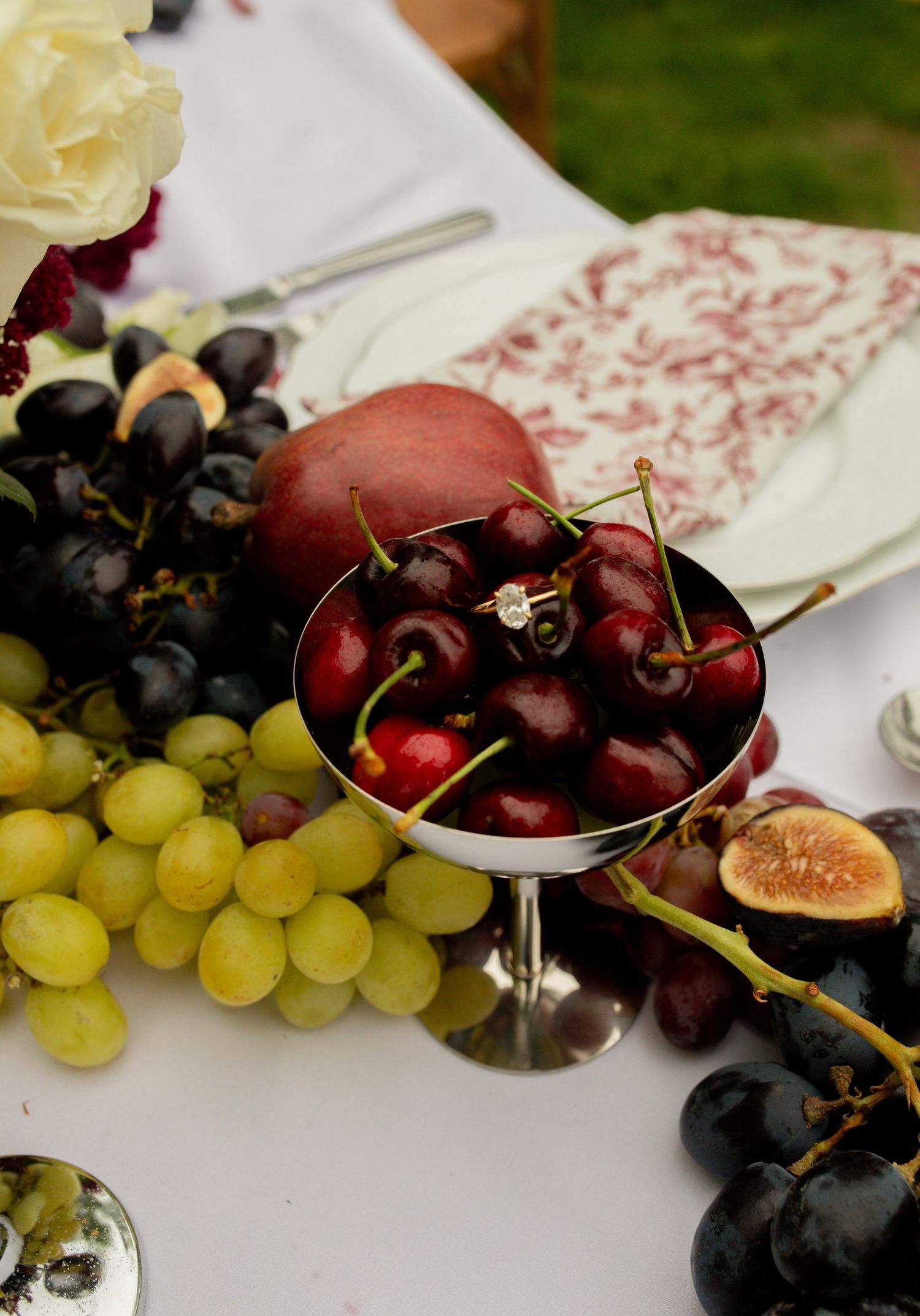 Silver bowl of fresh cherries with a ring, surrounded by green and black grapes, figs, and a peach on a white table.
