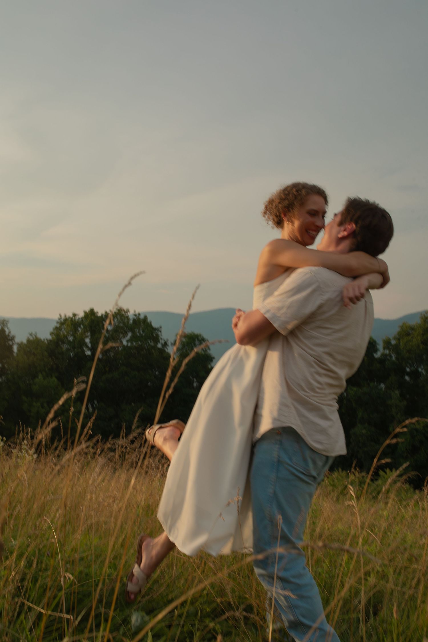 A man lifts a woman in a white dress in a golden meadow at sunset, sharing a romantic embrace.