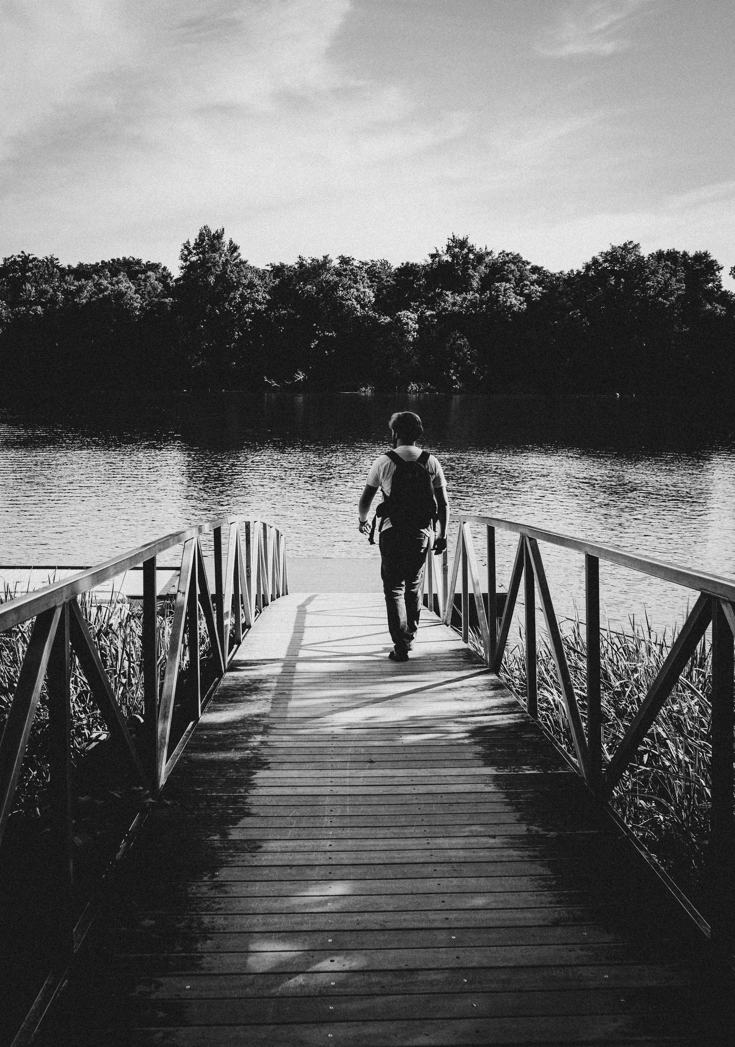 Black and white photo of person walking on wooden dock over serene lake with trees in background.