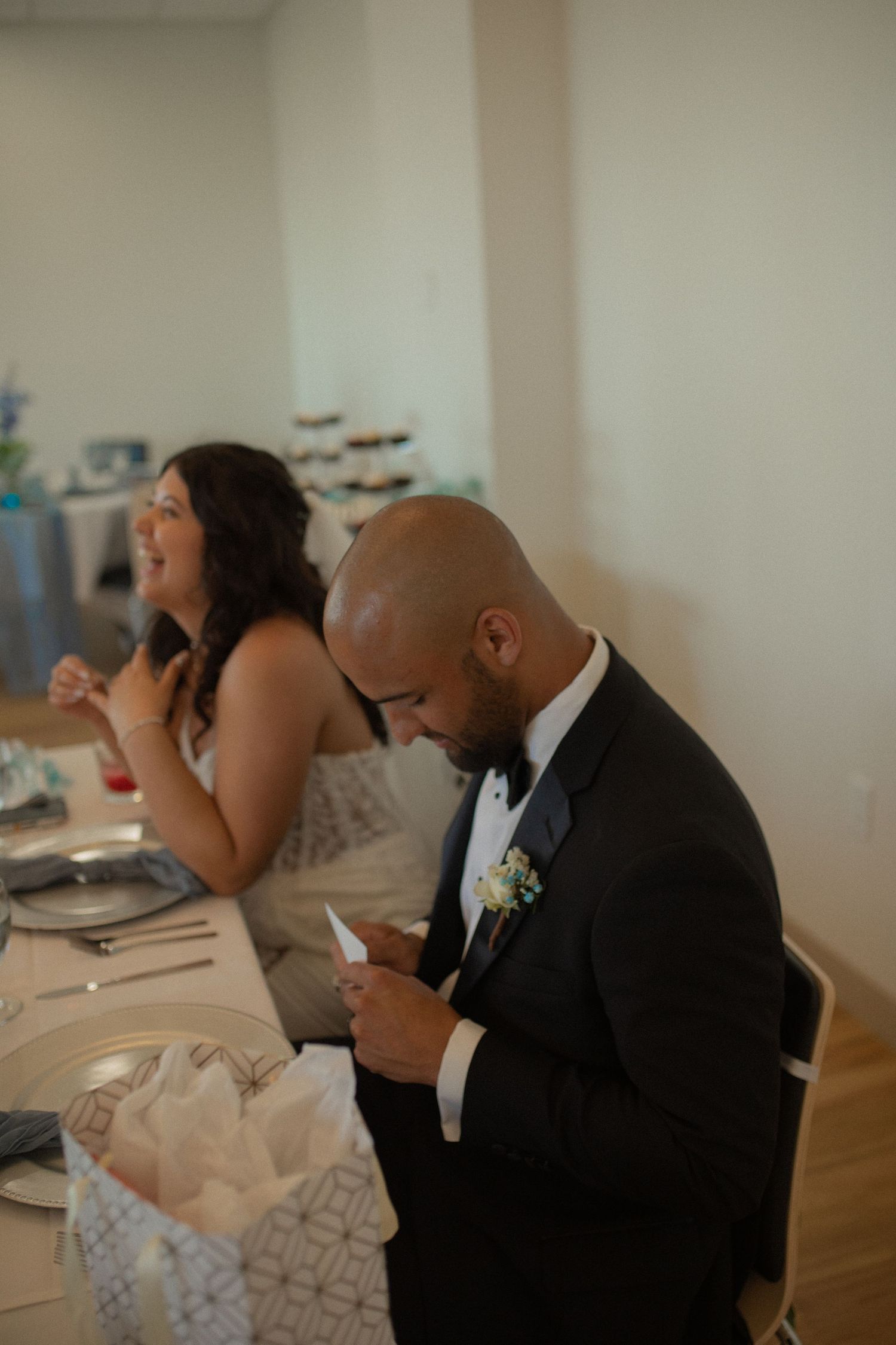 Groom in black tuxedo reads a note at wedding reception table while bridesmaid laughs in background.