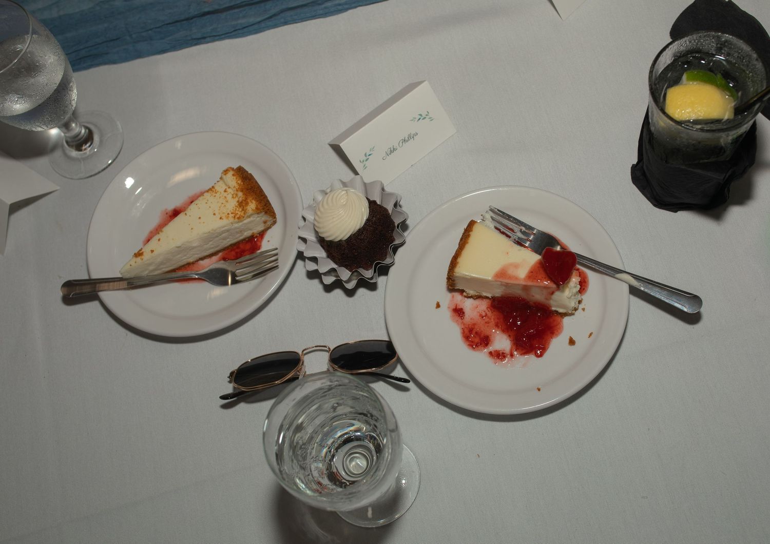 Three dessert plates with cheesecake slices and a cupcake on a white table, alongside sunglasses and drinks.