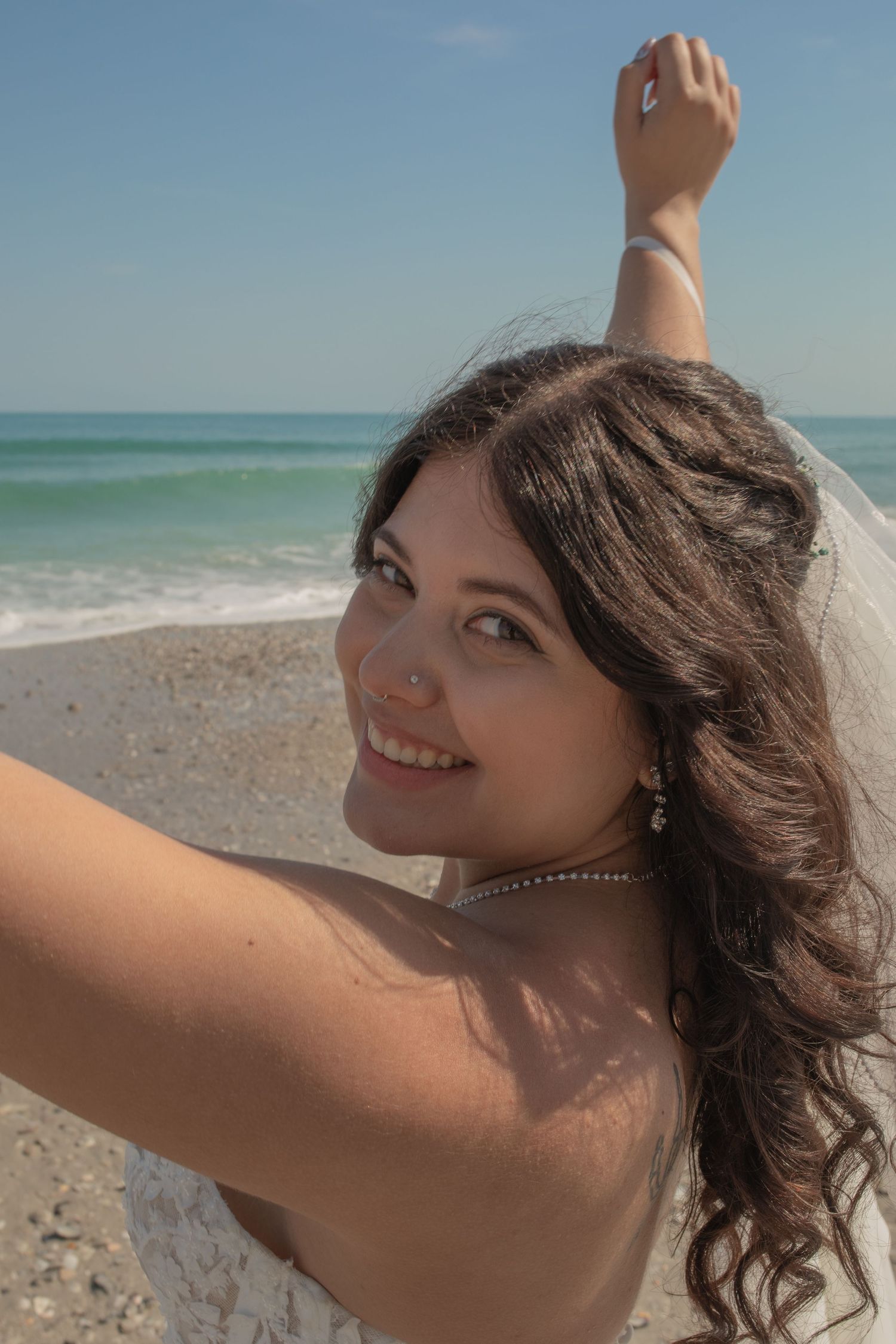 Smiling bride with dark curly hair holding veil at beach, wearing pearl necklace and earrings on sunny day.