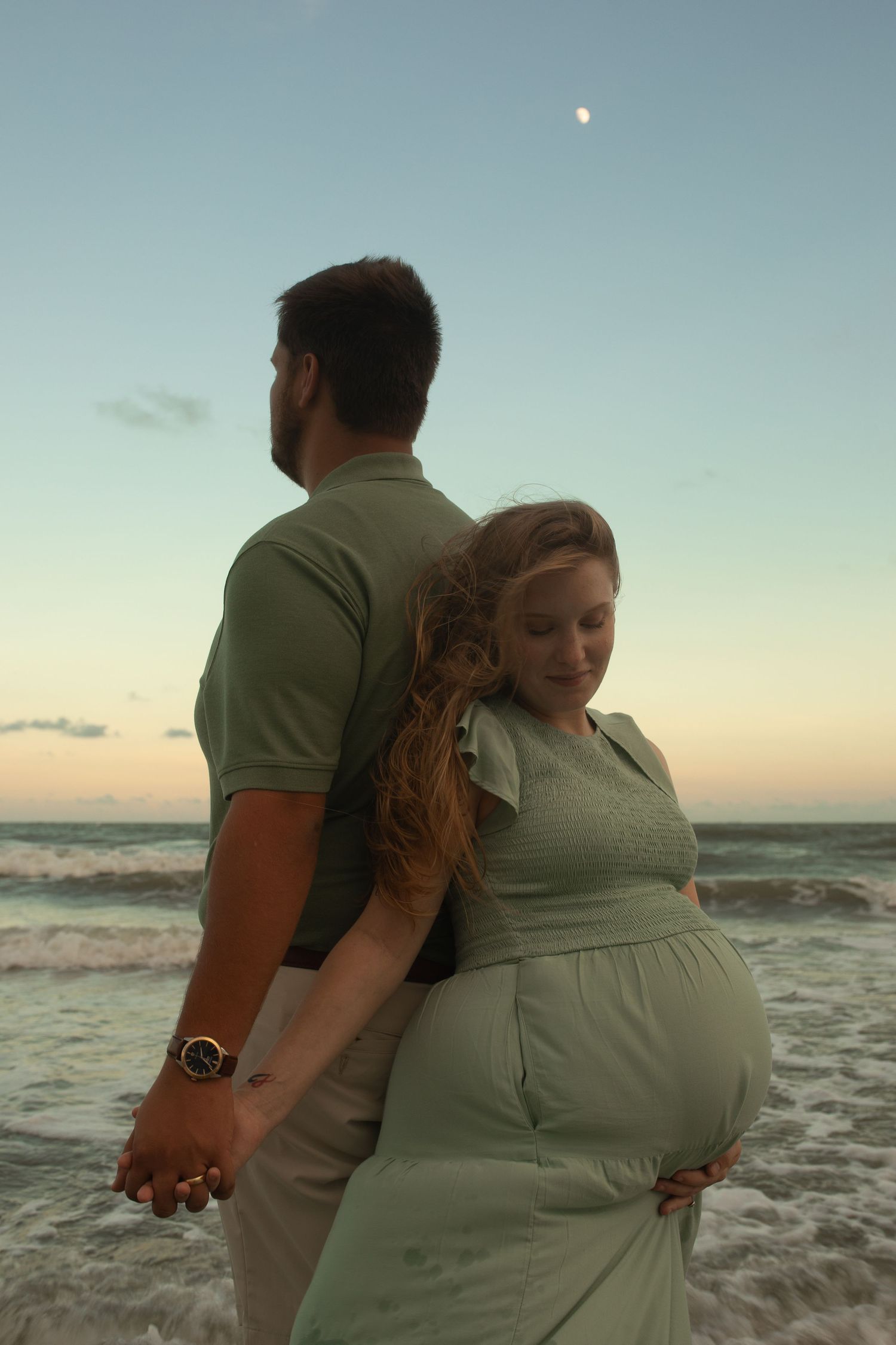 A couple in matching sage green outfits holds hands on the beach at sunset with the moon visible in the sky.