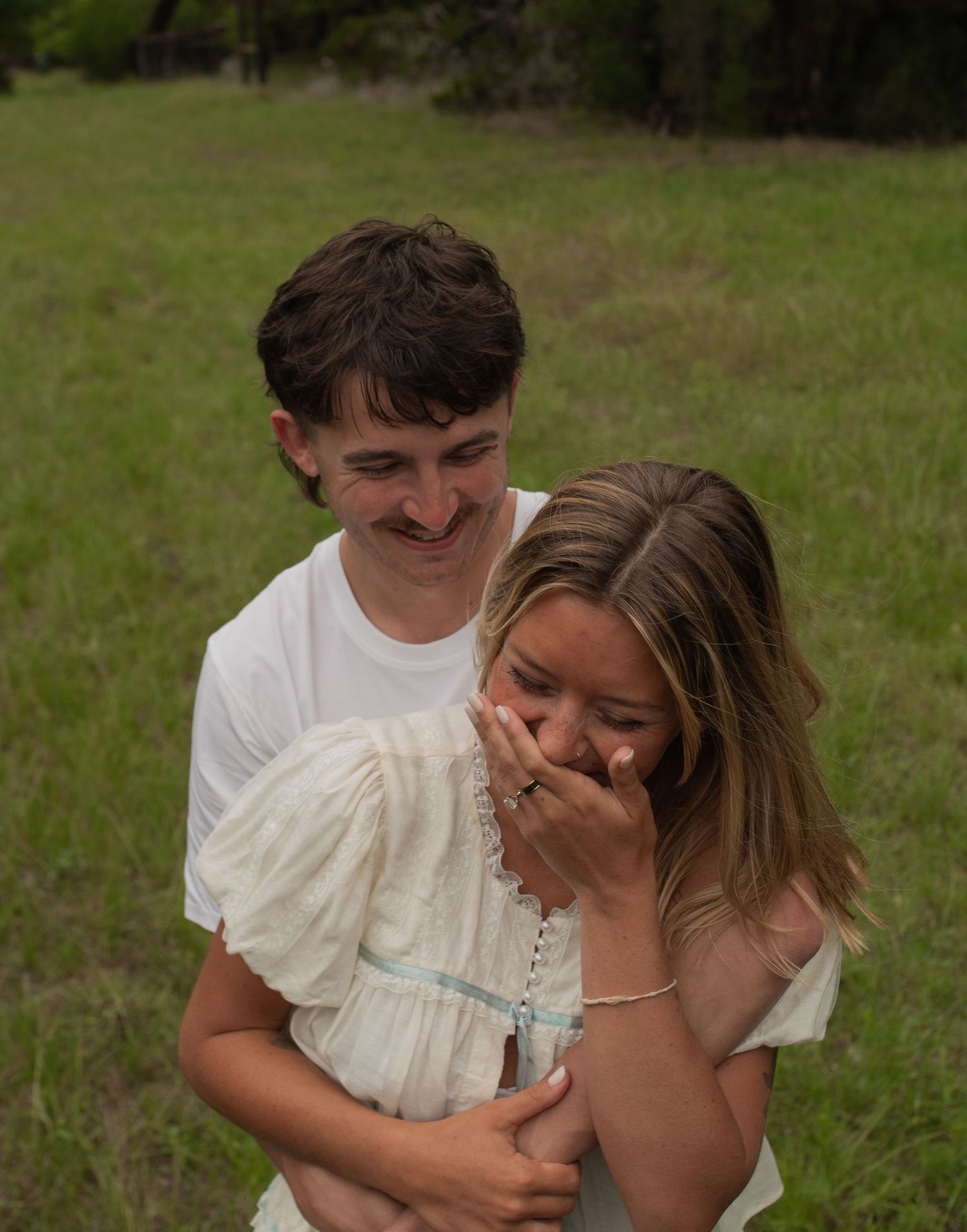 A young couple laughing and embracing in a green meadow, wearing white outfits during an outdoor photo session.