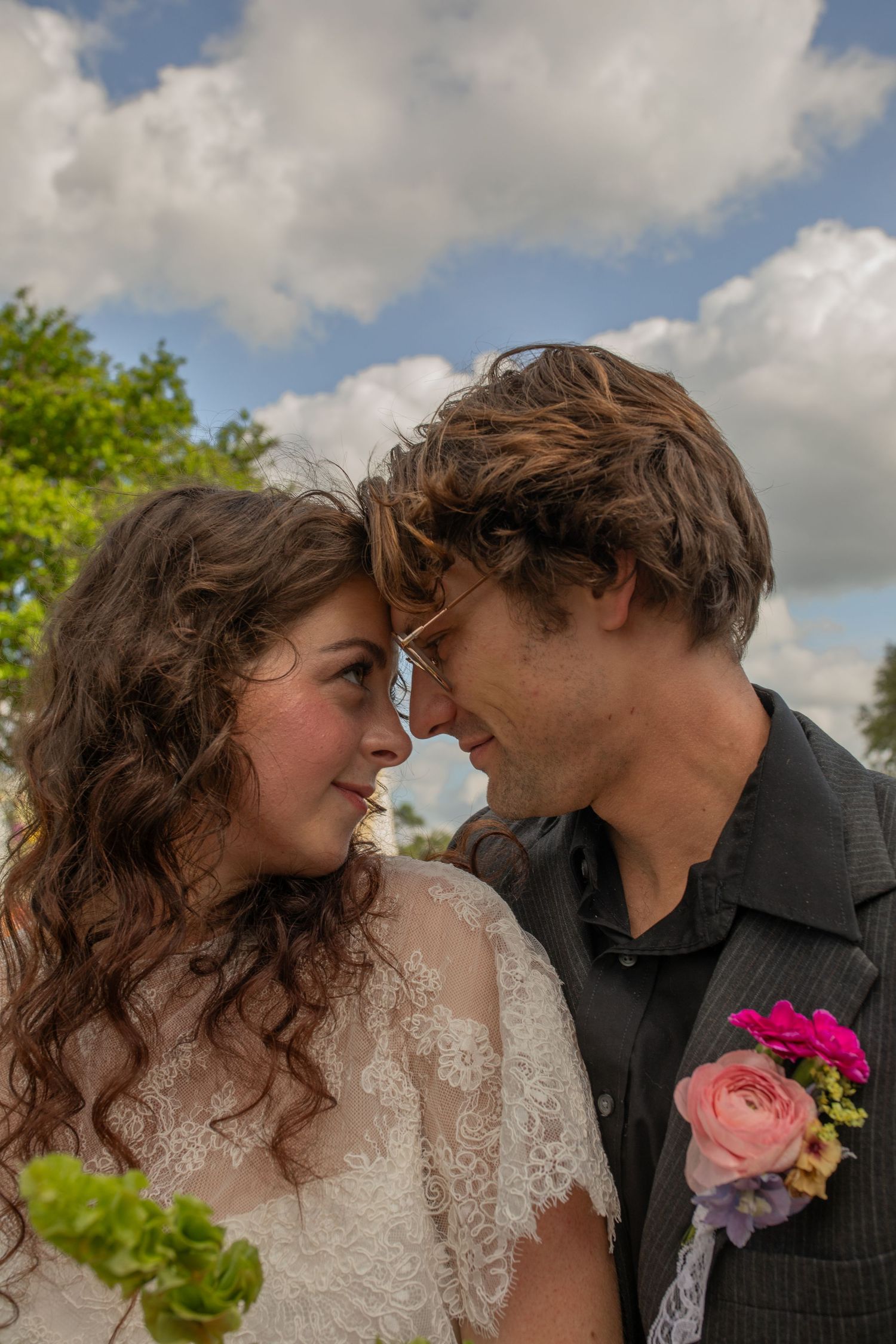 A bride and groom in wedding attire touch foreheads romantically outdoors under a partly cloudy blue sky.