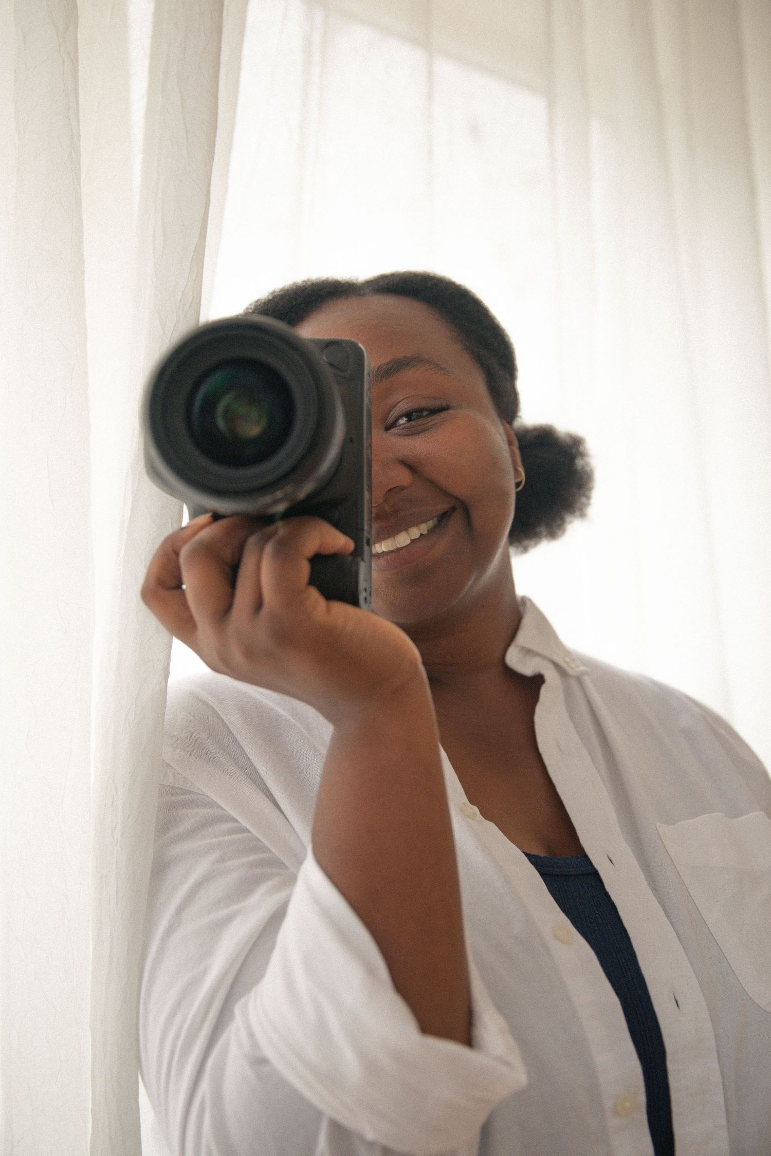 Smiling Black woman holding a DSLR camera up to her face, wearing a white shirt, standing near sheer white curtains.