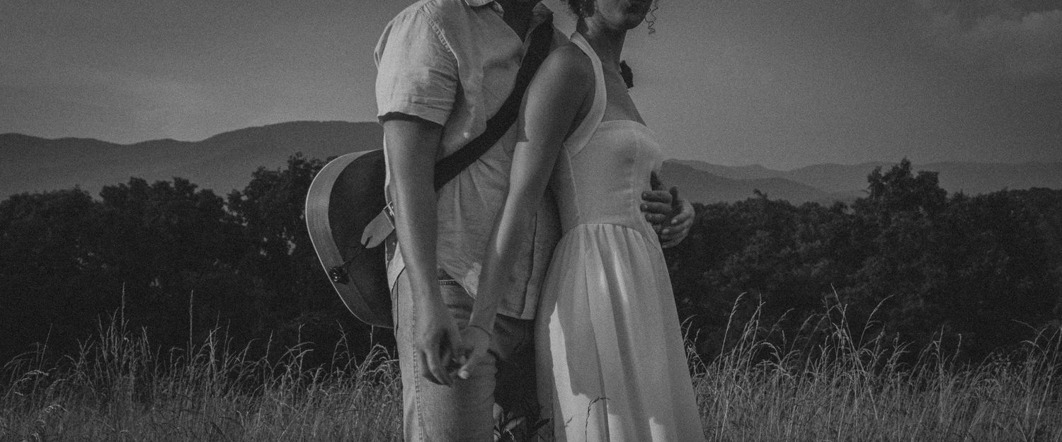 Black and white photo of a romantic couple holding hands in a field with mountains in the background.
