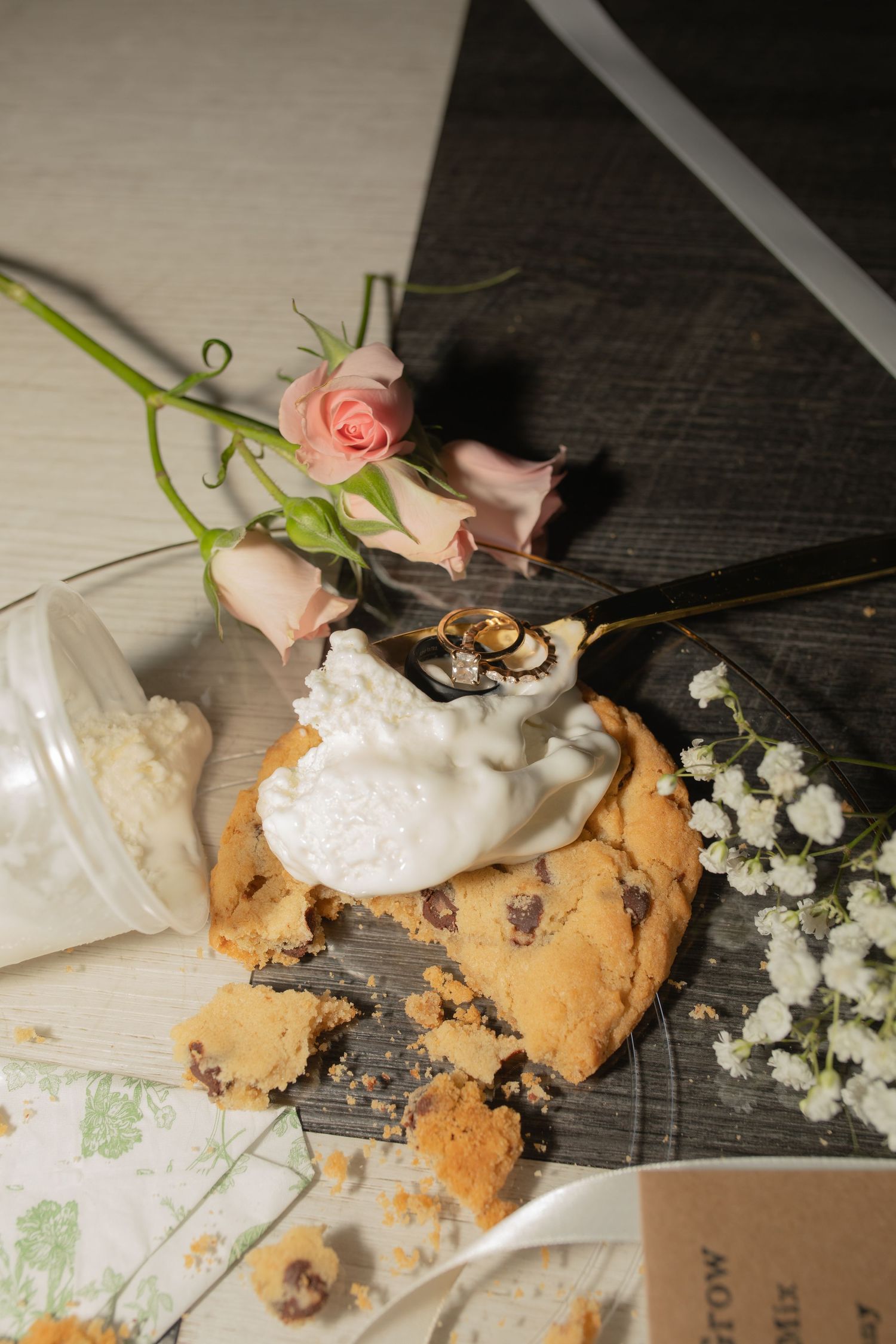 Wedding rings resting on a cream-topped chocolate chip cookie, surrounded by pink roses and baby's breath on a rustic wooden surface.