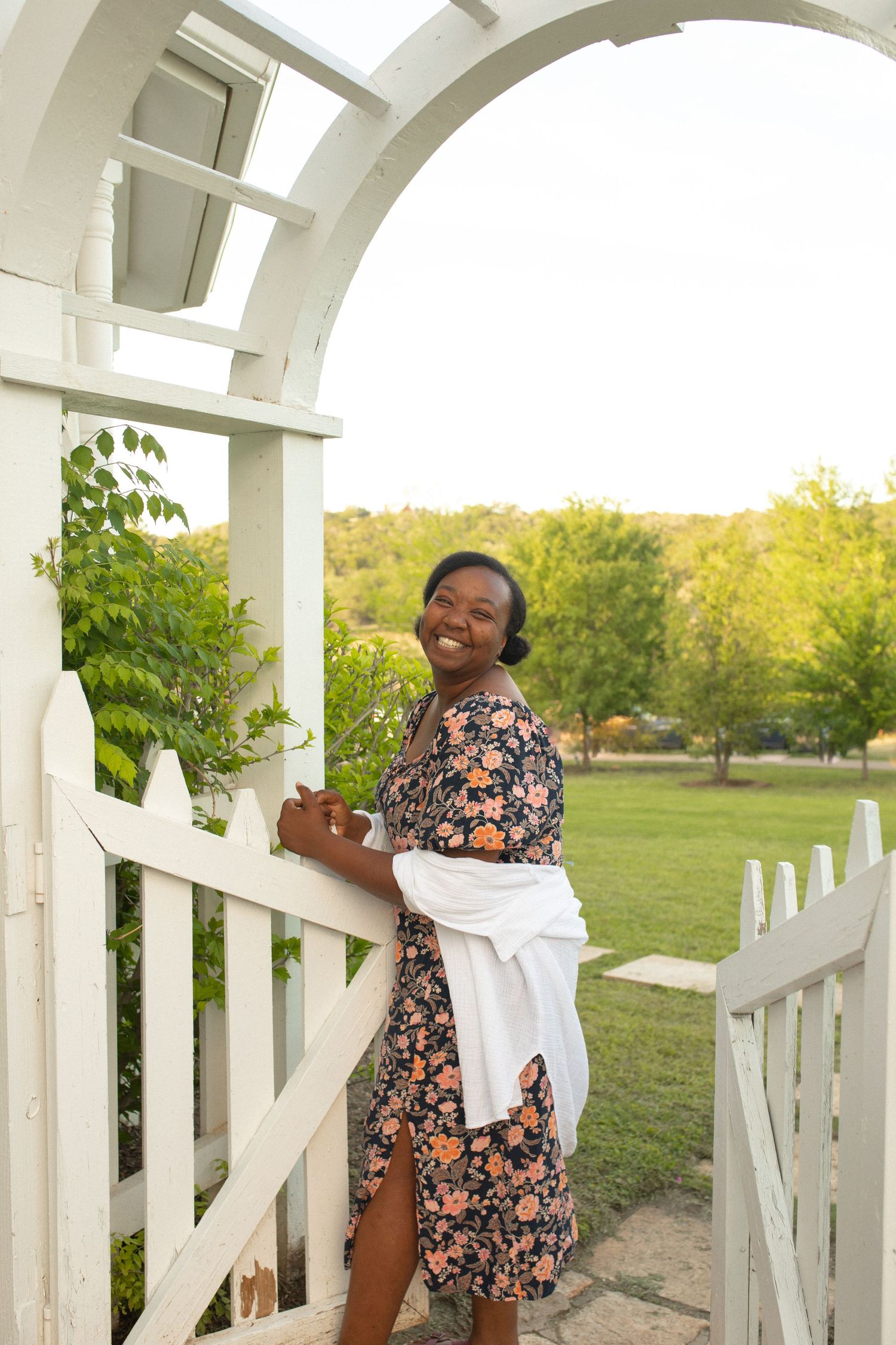 Smiling woman in floral dress standing at white garden gate arbor surrounded by lush greenery on sunny day.
