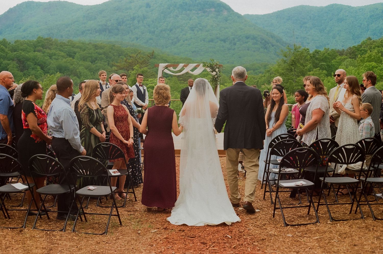 Outdoor wedding ceremony with bride and groom walking down the aisle, surrounded by guests and scenic mountain views.
