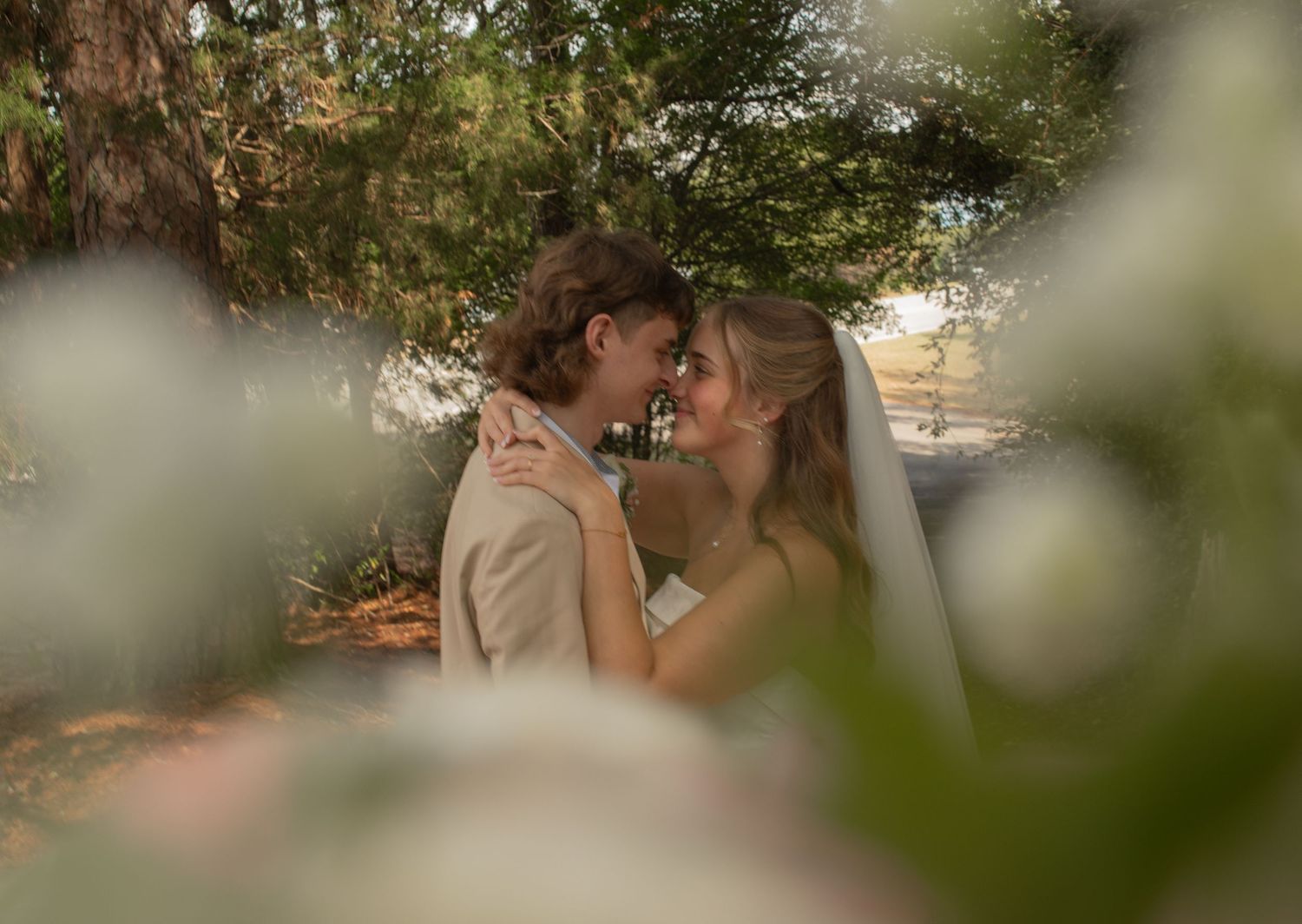 Romantic wedding couple sharing an intimate moment surrounded by soft white flowers and lush green trees outdoors.