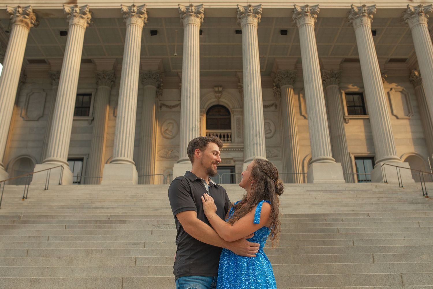 Couple embracing and smiling on steps of a grand columned building during golden hour.