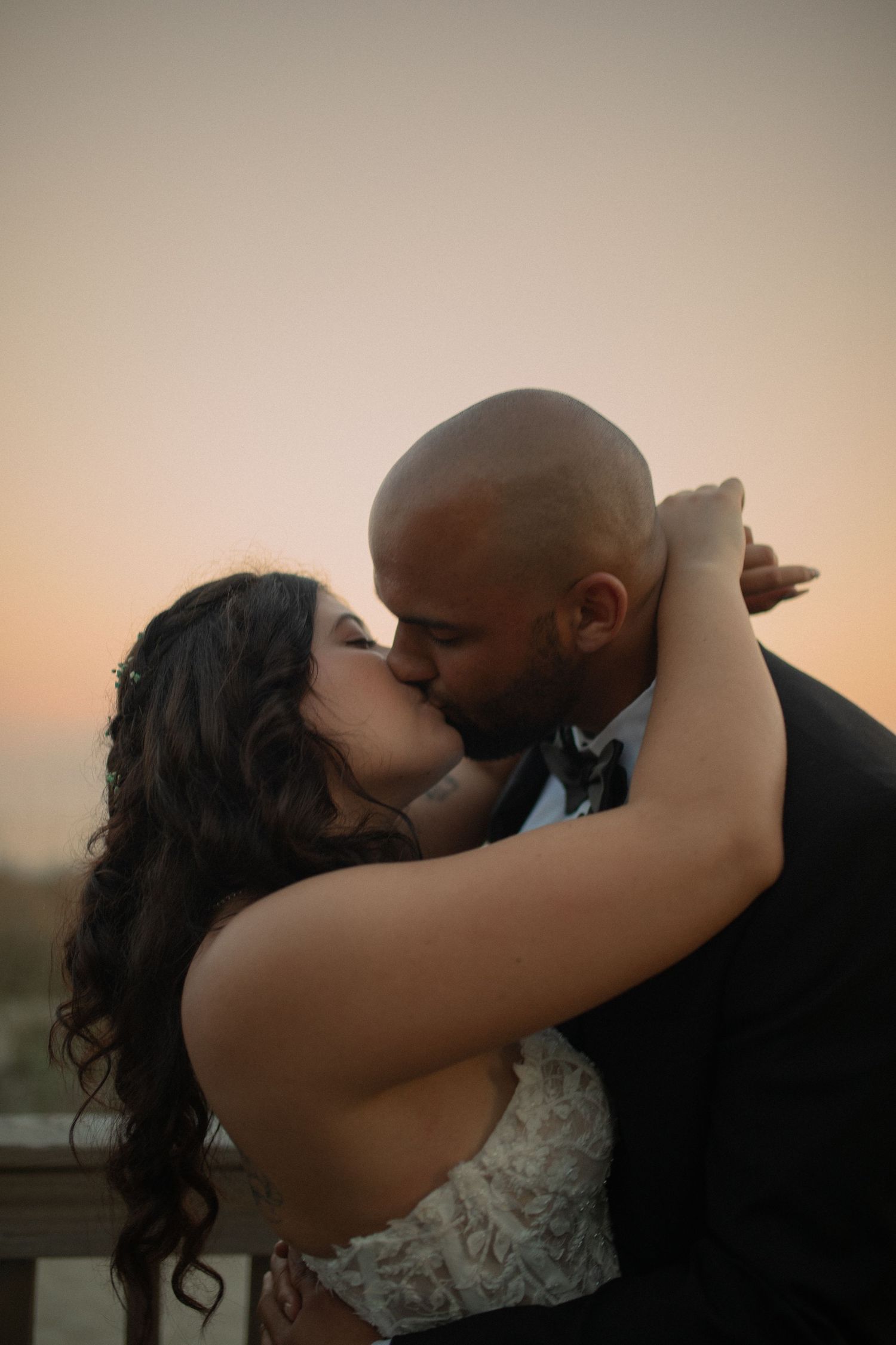 A bride and groom share a romantic kiss at sunset, dressed in wedding attire on an outdoor balcony.