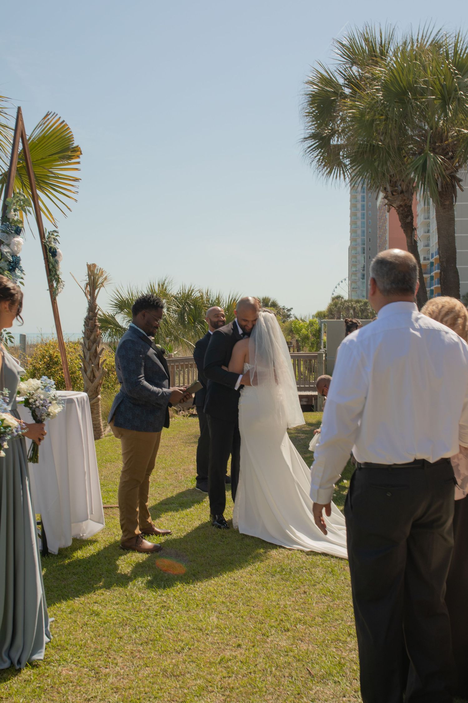 Outdoor beach wedding ceremony with bride and groom exchanging vows near palm trees and ocean backdrop on sunny day.
