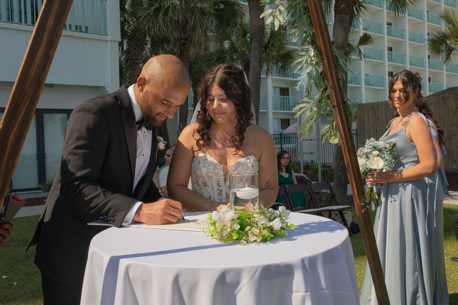 Bride and groom signing wedding documents at outdoor ceremony with bridesmaid holding bouquet in background.