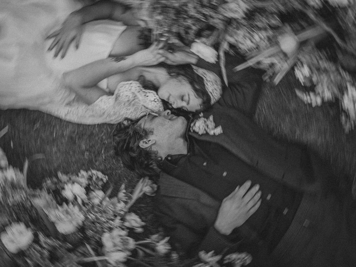 Black and white aerial photo of bride and groom lying on grass, kissing, surrounded by flower petals.
