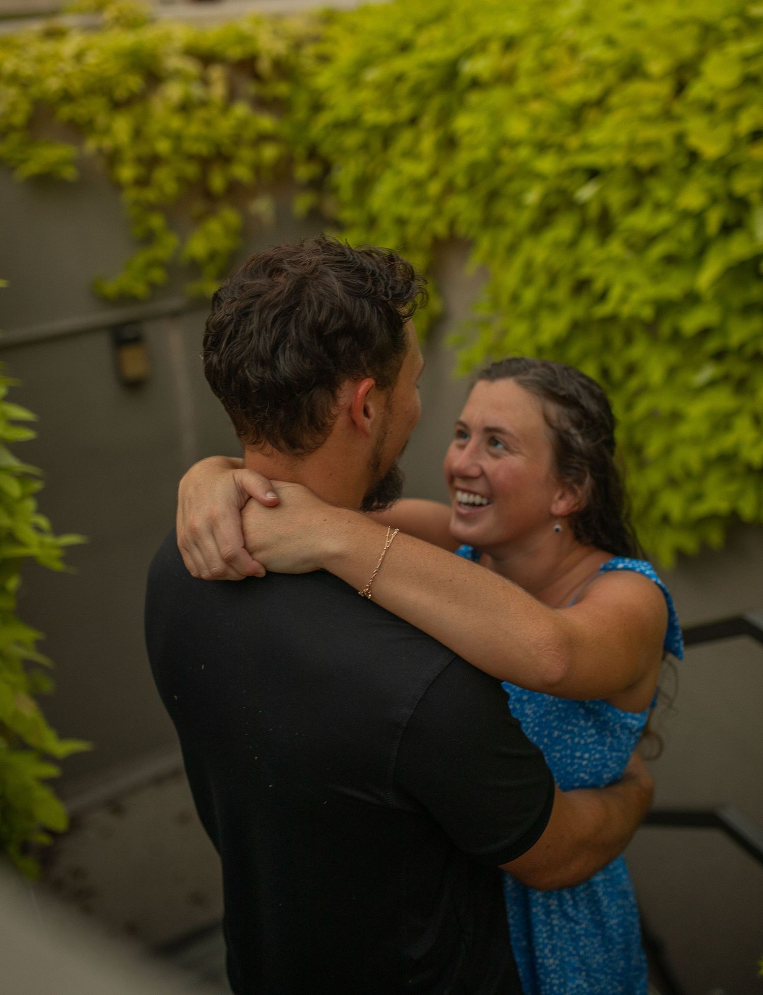 A smiling woman in a blue dress embraces a man in a black shirt outdoors, with lush green ivy in the background.