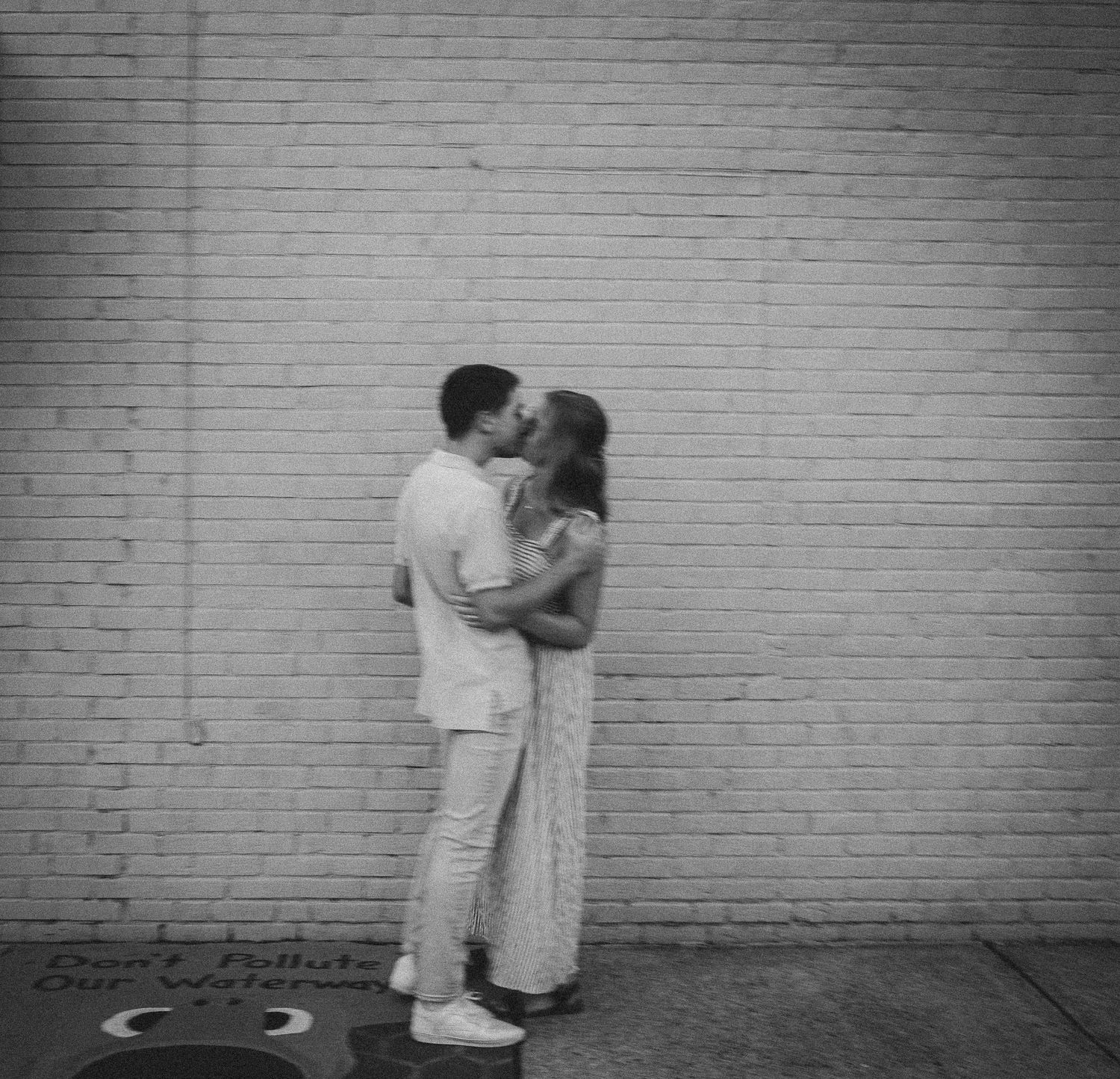 A romantic black and white photo of a couple embracing against a brick wall.